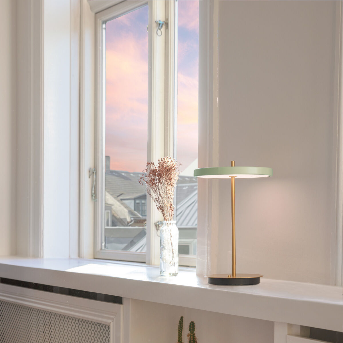 Tischlampe »Asteria Move II« auf einer weißen Fensterbank neben einem Fenster mit Blick auf den Sonnenuntergang. Die Lampe hat ein minimalistisches Design mit einer goldenen Stange und einem runden weißen Schirm. Auf der Fensterbank steht auch eine Dekovase mit getrockneten Blumen. Darunter befindet sich ein offenes Regal mit Büchern und einer kleinen Vase.