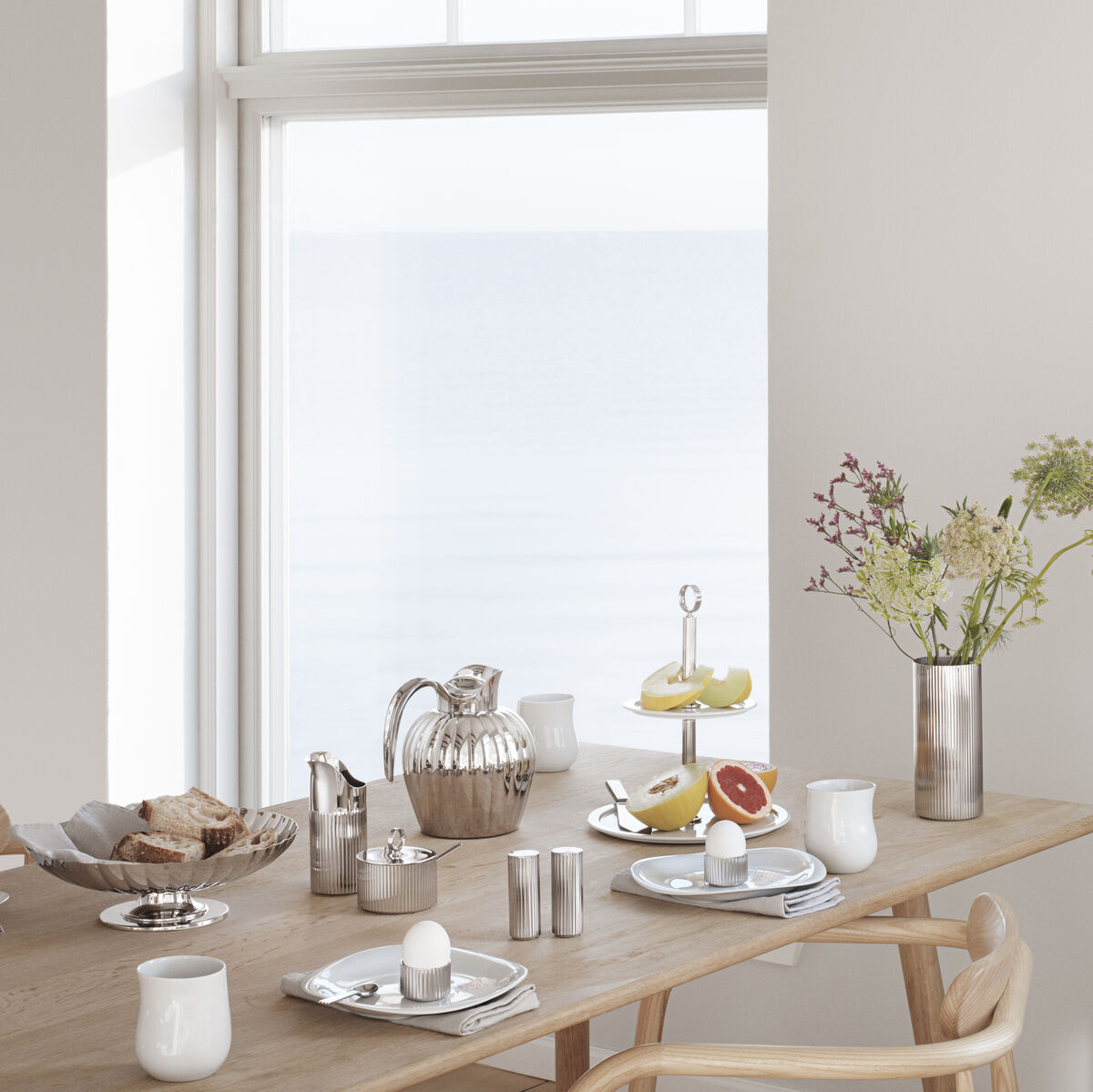 Ein elegant gedeckter Frühstückstisch mit weißen Wänden und großen Fenstern im Hintergrund. Der Tisch ist mit einem Salz- und Pfefferstreuer aus Edelstahl, einer Kaffeekanne, Tassen, einem Etagere mit Obst und einer Vase mit Blumen dekoriert. Holzstuhl und warme, helle Farben erzeugen eine gemütliche Atmosphäre.