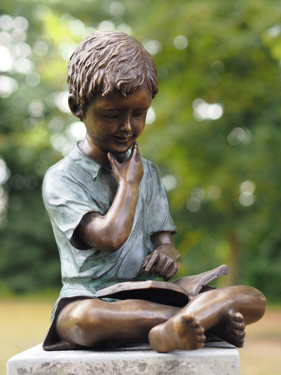 Eine Gartenskulptur zeigt einen sitzenden Jungen, der ein Buch liest. Die Skulptur ist in Braun- und Grüntönen gehalten und steht im Freien vor einem Hintergrund aus grünem Laubwerk.