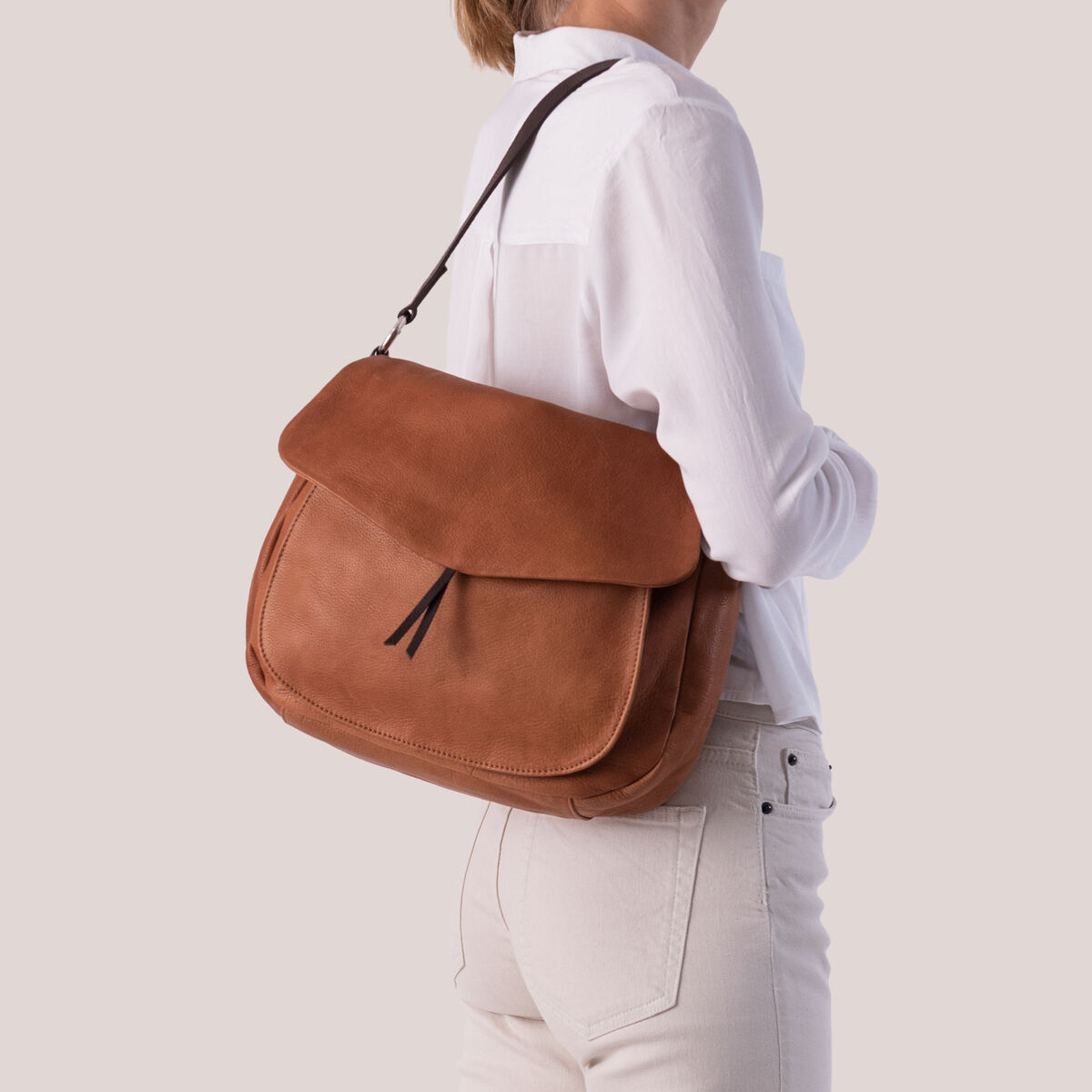 Eine Frau trägt eine braune Lederslingbag über ihrer Schulter. Sie trägt eine weiße Bluse und beige Hose. Die Tasche hat einen minimalistisch und stilvoll gestalteten Look.