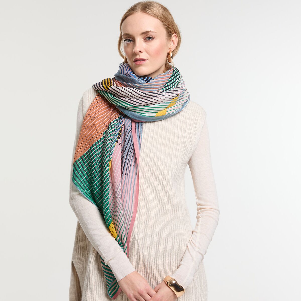 Eine Frau trägt einen großen, mehrfarbigen Wollschal mit Streifenmuster in Blau, Grün, Gelb, Rosa, Orange, Weiß und Schwarz. Der Schal ist elegant um den Hals gelegt und bildet einen Kontrast zum hellen Kleid der Frau. Das Bild eignet sich zur Darstellung moderner, gemütlicher und vielfältiger Damenmode.