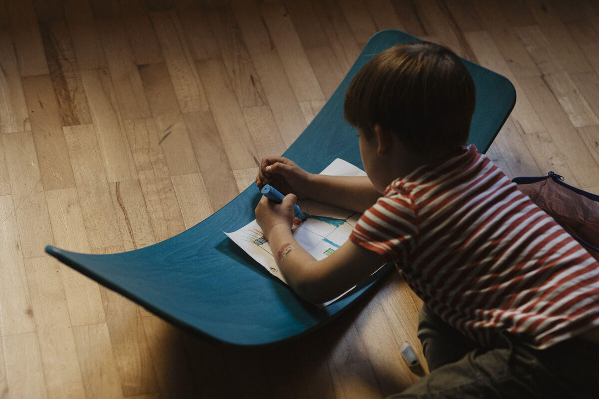 Ein Kind liegt auf einem mintgrünen Balance Board 'das.Brett' und malt auf einem Blatt Papier. Der Fußboden ist aus Holz.