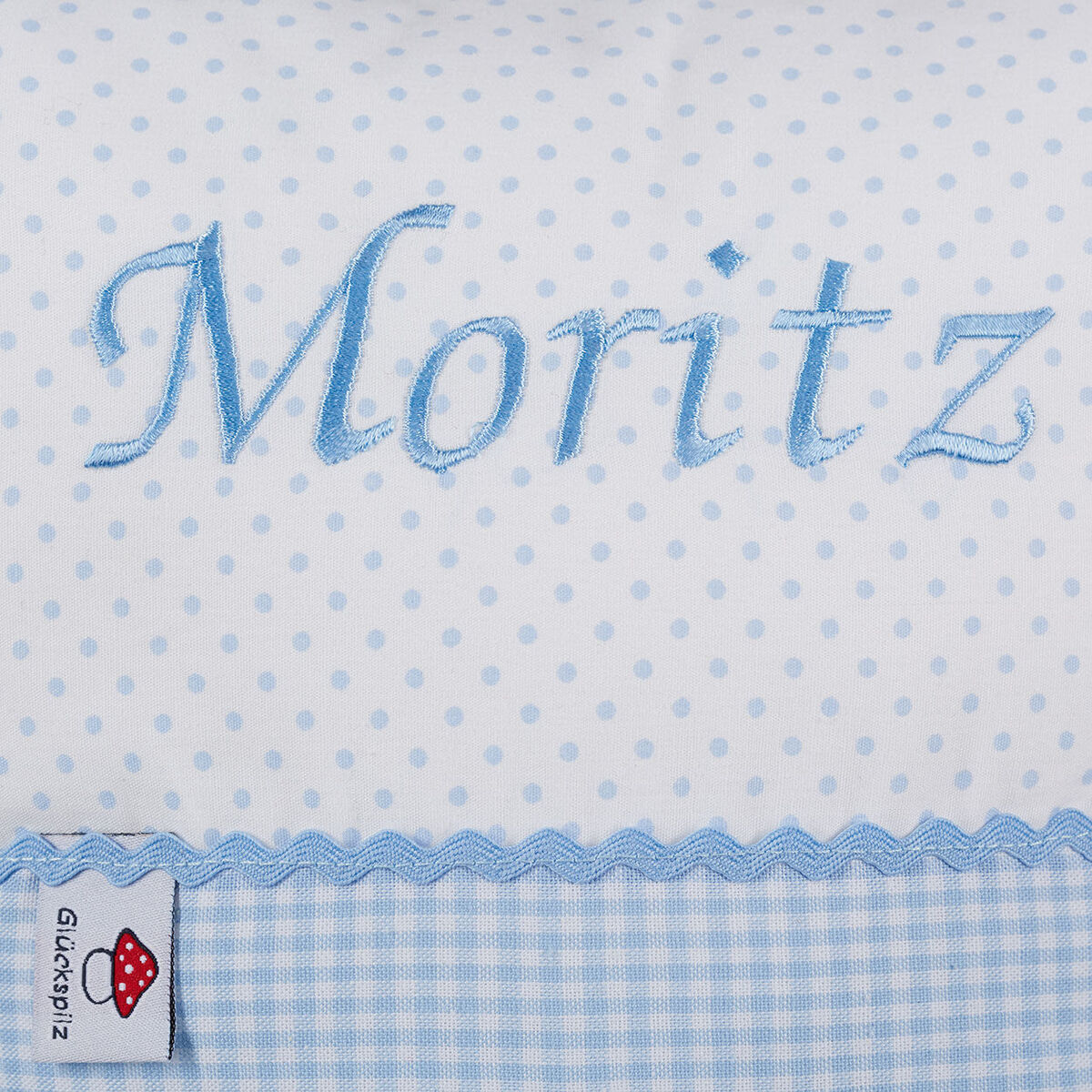 Ein personalisiertes Kissen mit dem Namen „Moritz“, gestaltet in hellblauer Schrift mit Polka-Dots-Hintergrund in Weiß und Blau. Das Kissen hat eine Rüscheneinfassung und ein kariertes Muster am unteren Rand.