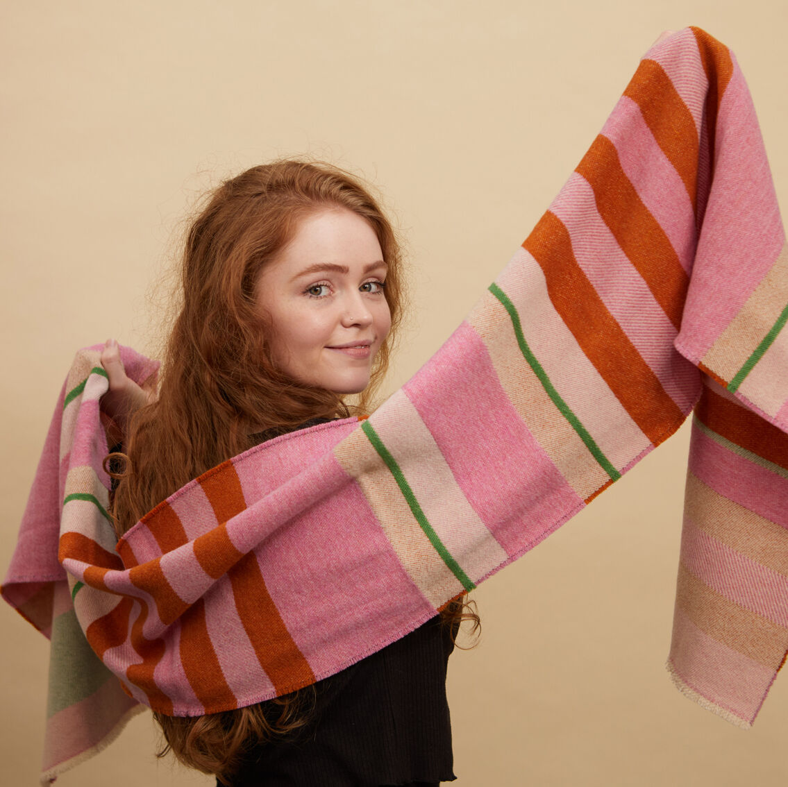 Eine Frau trägt einen Schal mit dem typischen Bauhaus Stölzl Design. Der Schal hat ein Streifenmuster in Rosa, Orange, Grün und Beige. Der Hintergrund ist hell.