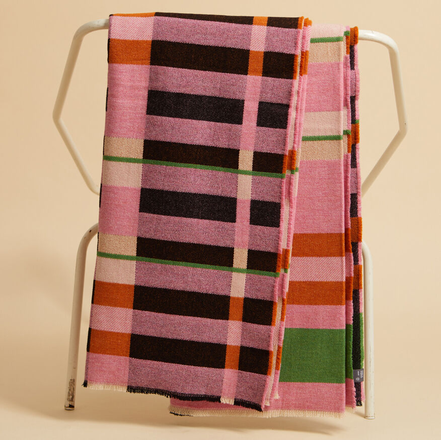 Eine farbenfrohe Decke mit gestreiften Mustern in Pink, Schwarz, Weiß, Grün und Orange, drapiert über ein minimalistisches drahtiges Gestell vor einem beigen Hintergrund.