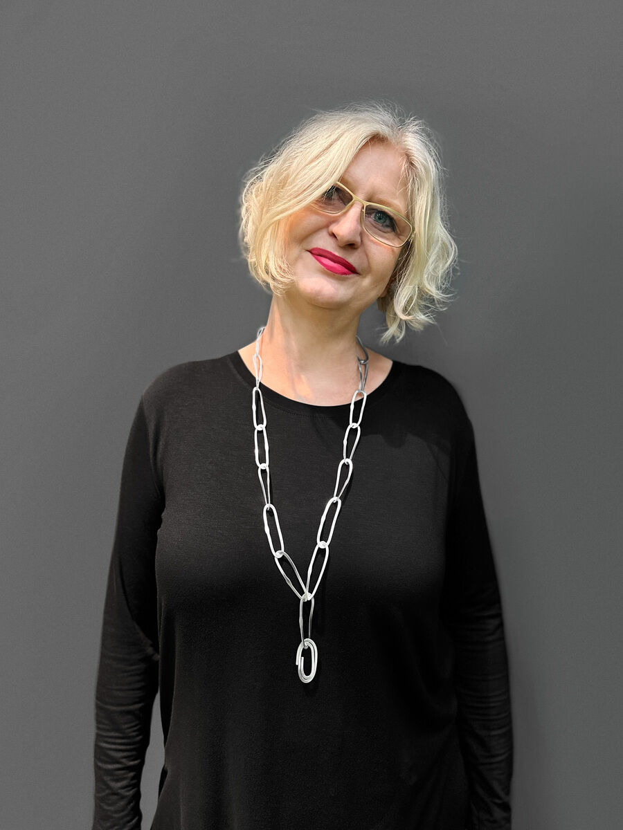 Dame mit blonder Frisur trägt eine lange silberne Halskette mit wellenförmigem Anhänger vor einem grauen Hintergrund, gekleidet in einem schwarzen Oberteil.