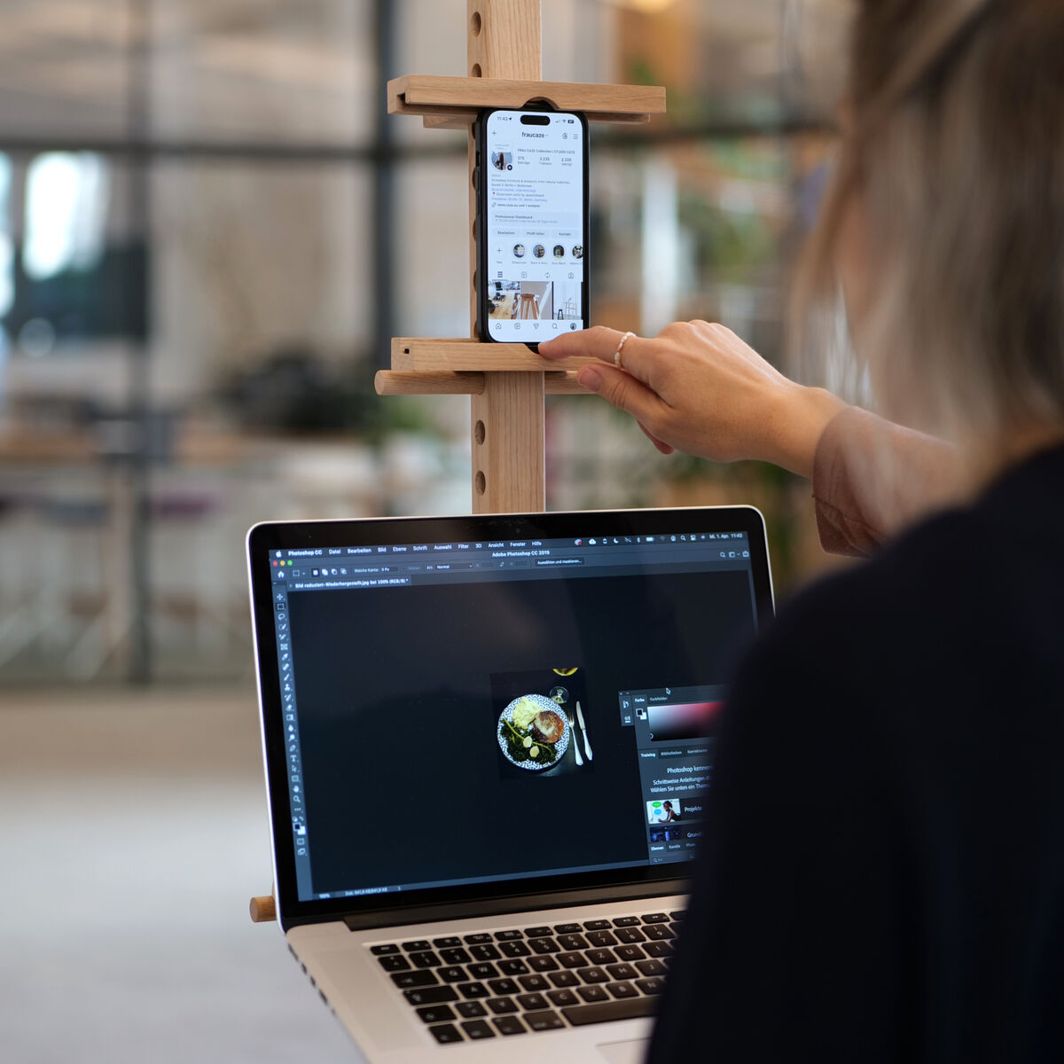 Ein mobiles Device Stativ aus naturbelassener Eiche und Birkensperrholz von Frau Caze steht in einem modernen Büroraum. Auf den verstellbaren Ablageflächen sind ein Smartphone und ein Laptop platziert. Eine Person arbeitet am Laptop und bedient das Smartphone.