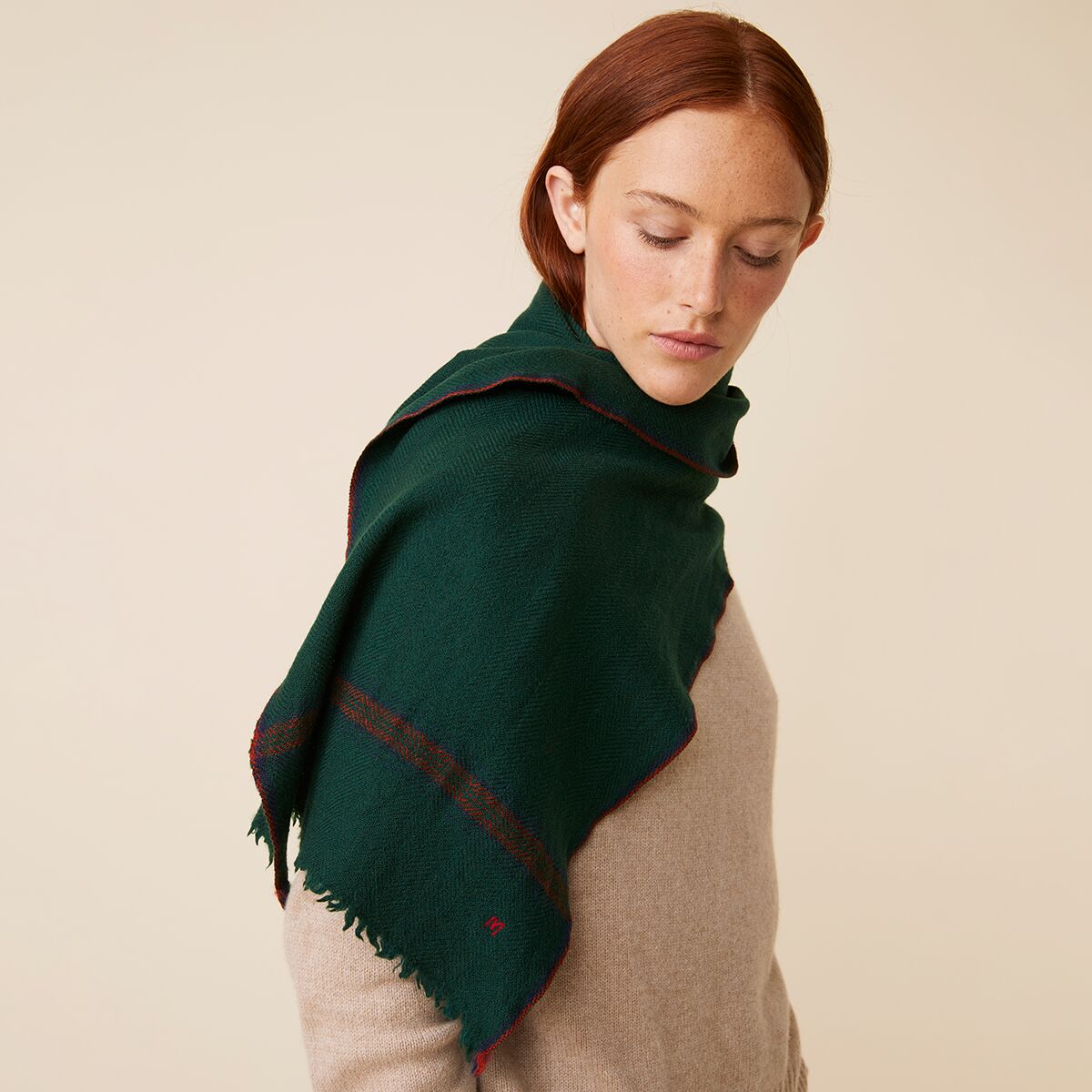 Frau mit einem dunkelgrünen, hochwertig verarbeiteten Schal aus 100% Schurwolle von Moismont. Der Schal ist handbedruckt, liegt locker über ihre Schultern und zeigt feine rote Streifen und Fransen an den Kanten. Sie trägt einen beigen Wollpullover und blickt ruhig zur Seite.