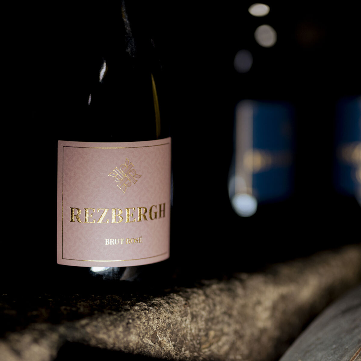 Eine edle Flasche Crémant de Luxembourg Brut Rosé mit einem eleganten, rosafarbenen Etikett steht im Vordergrund. Der Flaschenhals ist teilweise sichtbar, rechts im Bild erscheinen unscharf weitere Flaschen. Die Szene ist von dunklen, luxuriösen Tönen dominiert, die Präsentation wirkt exklusiv und feierlich.