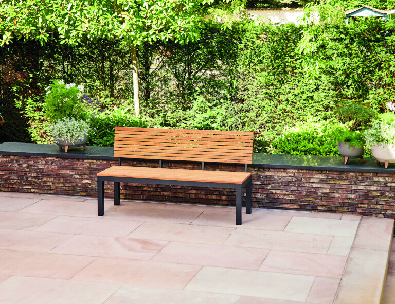 Eine Holzbank mit schwarzem Metallgestell steht auf einer Terrasse mit Steinboden, umgeben von einer Gartenmauer mit diversen Pflanzen im Hintergrund.