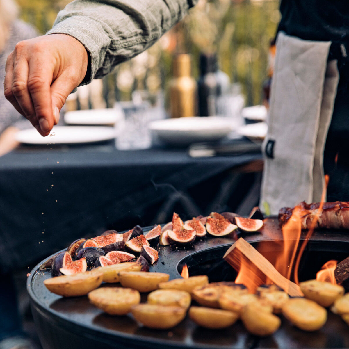 Eine hand hält eine Feige über einem Outdoor-Grill mit Feuerschale und Halbring, auf dem Kartoffeln und Feigen liegen. Im Hintergrund ist eine Tischszene mit Flaschen und Geschirr zu erkennen.