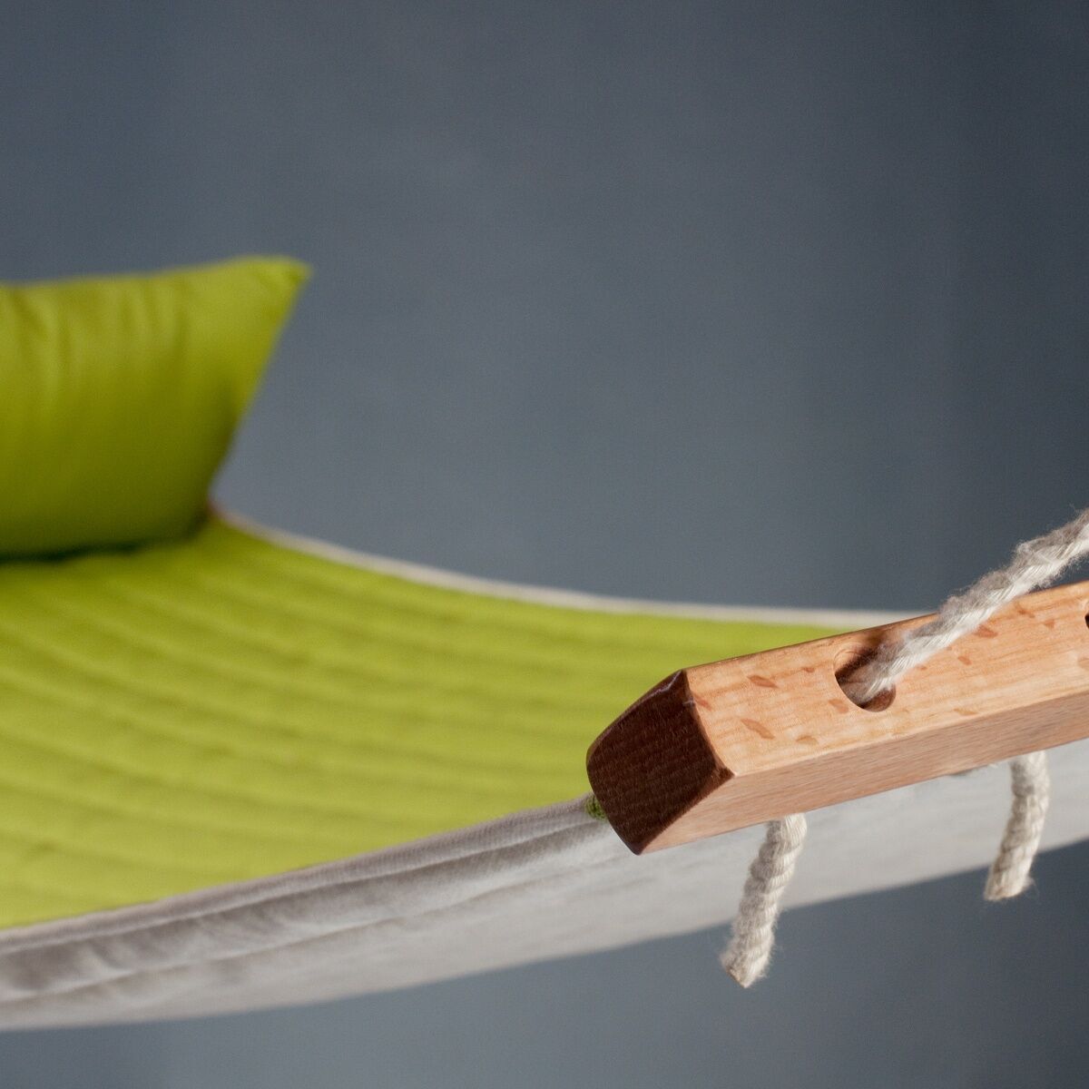 Eine grüne und weiße Hängematte »Alabama« aus Holz und Stoff, die vor einem blauen Hintergrund aufgehängt ist. Ein Kissen in passendem Grün liegt auf der Hängematte.