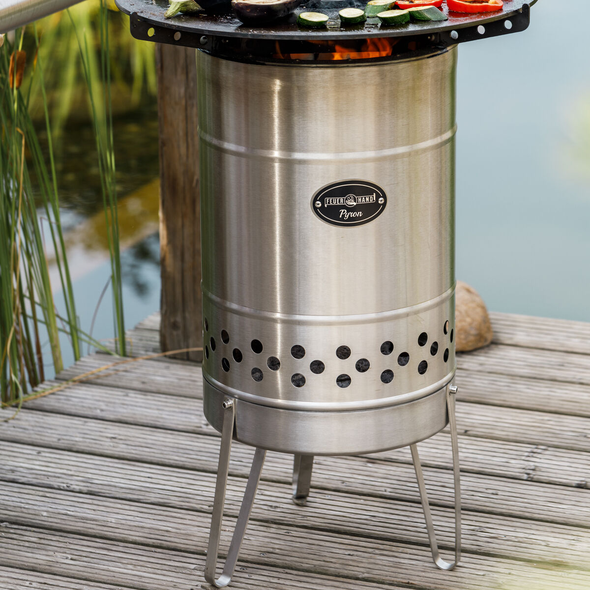 Eine Feuertonne aus Edelstahl der Marke »Pyron« steht auf einer Holzterrasse. Auf dem Grillrost oberhalb der Tonne befinden sich Scheiben von Zucchini und Paprika. Im Hintergrund ist ein Teich mit Schilf und Gras zu sehen, der eine entspannte Outdoor-Atmosphäre vermittelt.