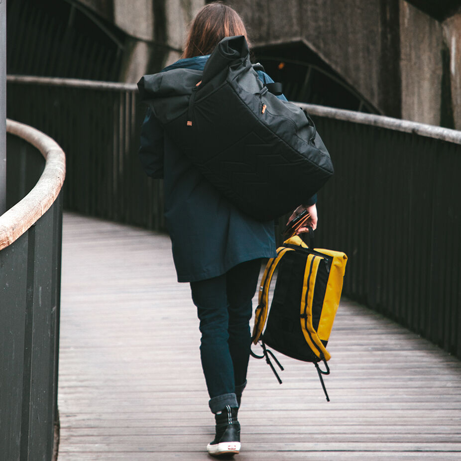 Eine Person geht auf einem Holzsteg und trägt eine große schwarze Duffle Bag »Smögen« auf der Schulter. In der linken Hand hält sie einen gelb-schwarzen Rucksack. Die Person ist mit einer dunkelblauen Jacke und einer dunklen Hose bekleidet.