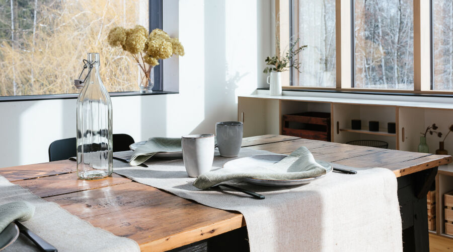 Holztisch mit hellen Stoffservietten und zwei grauen Tassen in einem hellen Esszimmer mit großen Fenstern