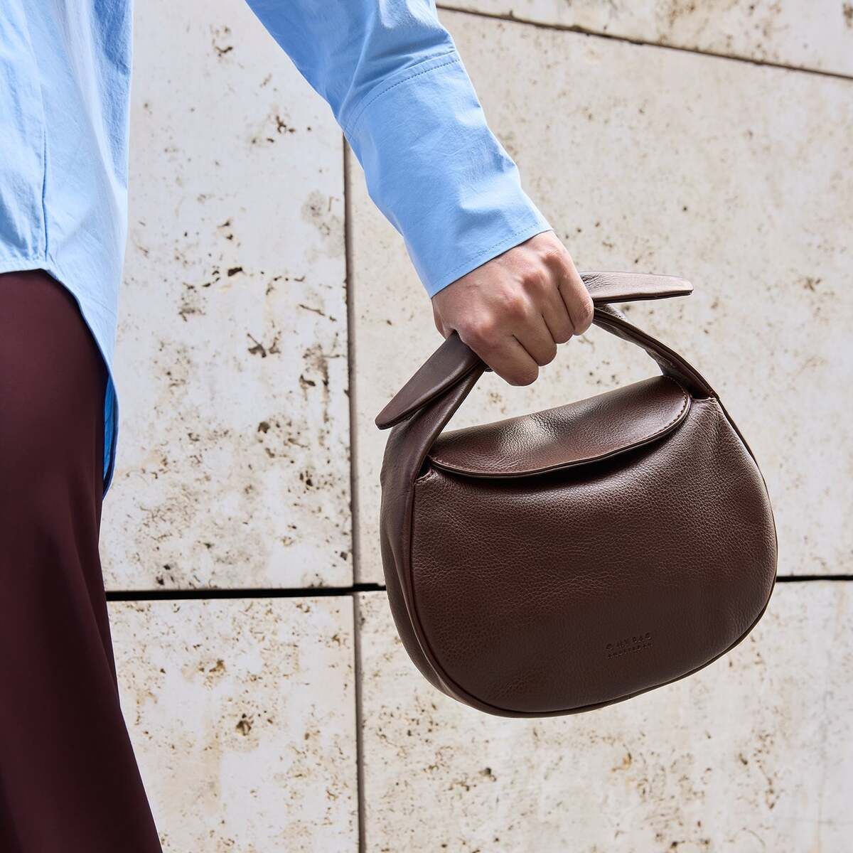 Eine Person hält eine braune, halbmondförmige Tasche namens 'Coco Croissant' aus Leder in der Hand. Die Person trägt ein hellblaues Hemd und einen dunkelbraunen Rock. Im Hintergrund ist eine beige Mauer mit leichten Sprenkeln zu sehen.