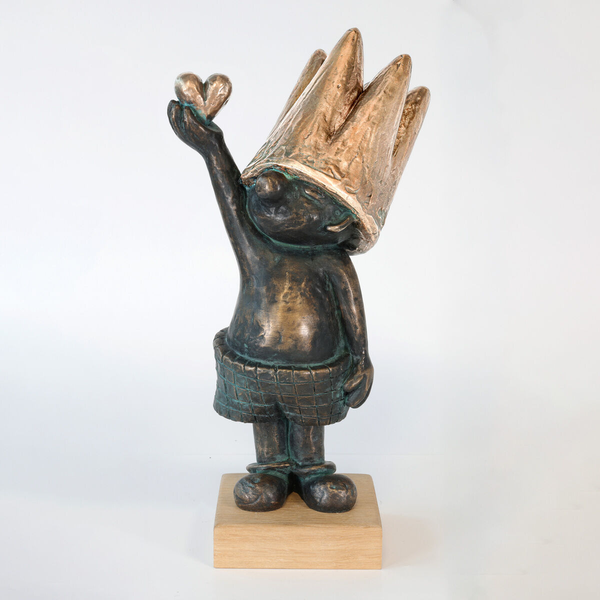 Die Bronzeskulptur »Guck maaal… für Dich!« von Toby Wichmann zeigt eine humorvoll gestaltete Figur mit großer, goldener Krone, erhobenem Arm und Herz in der Hand. Auf einem eckigen Holzsockel montiert. Oberfläche teils poliert und patiniert.