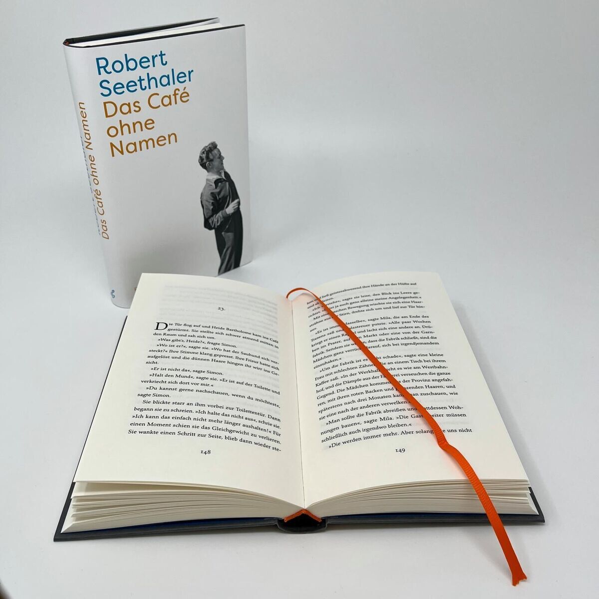 Auf dem Bild sieht man das Buch 'Das Café ohne Namen' von Robert Seethaler. Es ist ein Hardcover-Buch mit einem weißen Umschlag und schwarzem Text. Das Buch ist geöffnet und ein orangefarbenes Lesezeichen ist eingeklemmt.