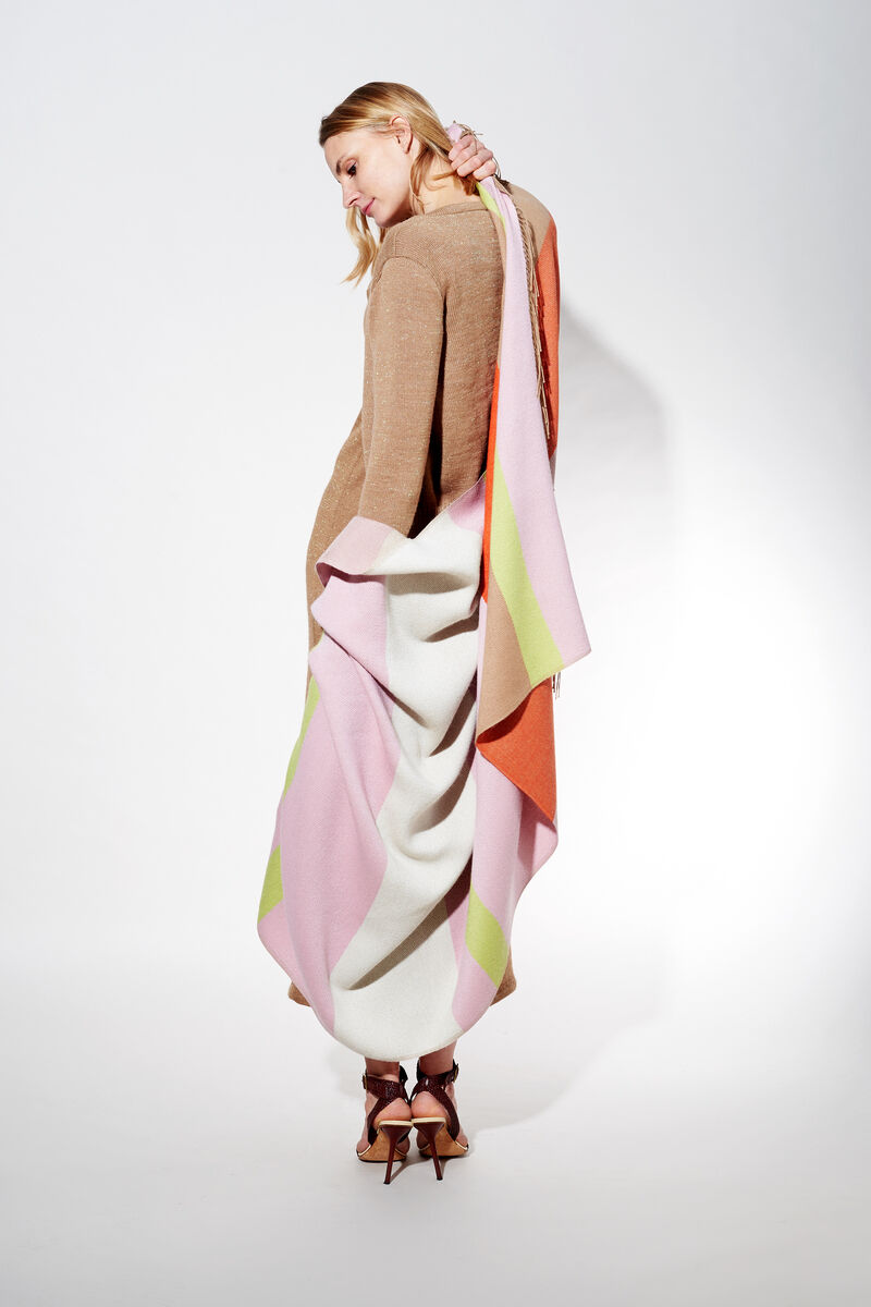 Blonde Frau trägt ein Cape aus Alpaka-Wolle in den Farben Rosa, Weiß, Grün und Orange. Die Kleidung scheint elegant und komfortabel.