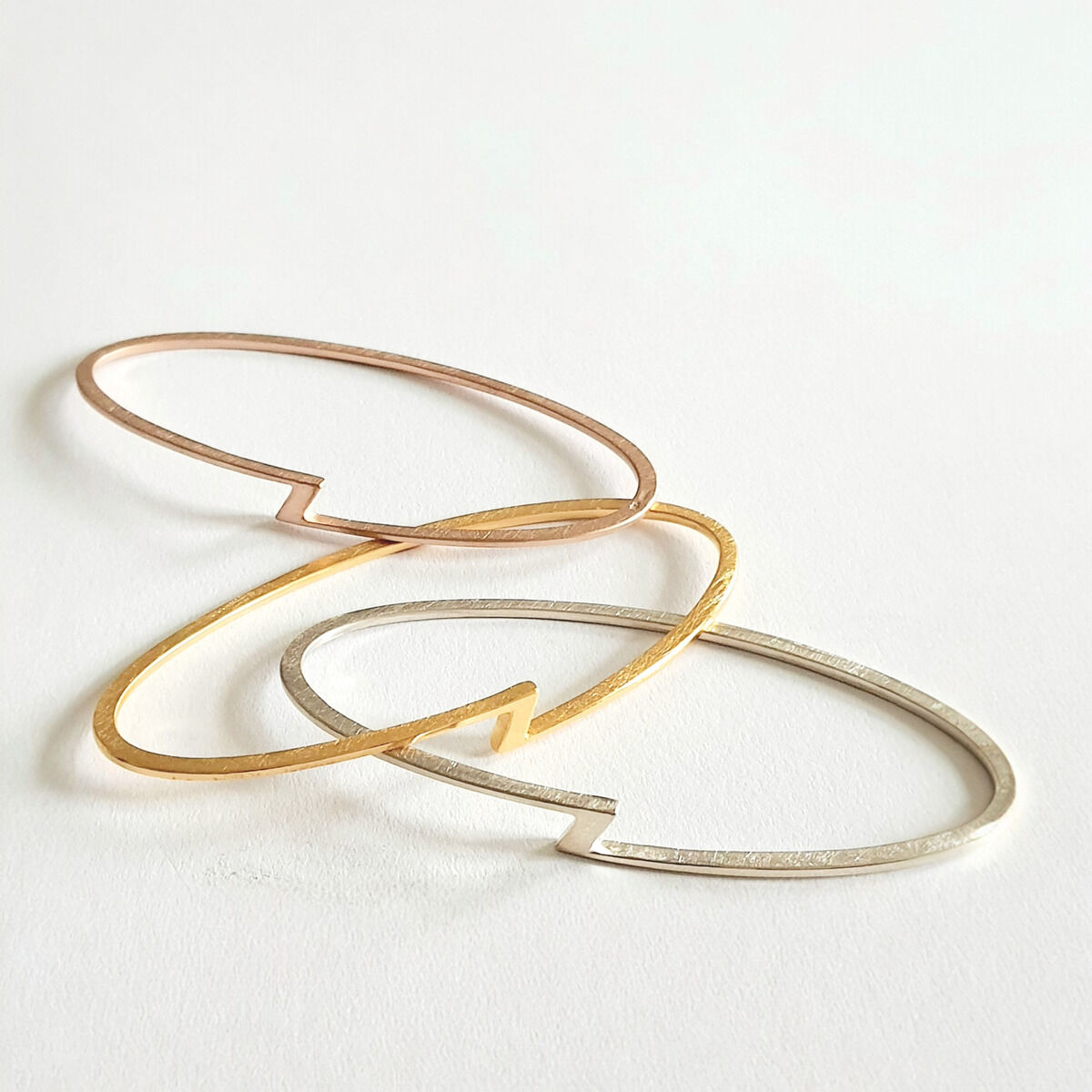 Drei schlichte, metallische Armreifen in Silber, Gold und Roségold liegen auf einer weißen Oberfläche.