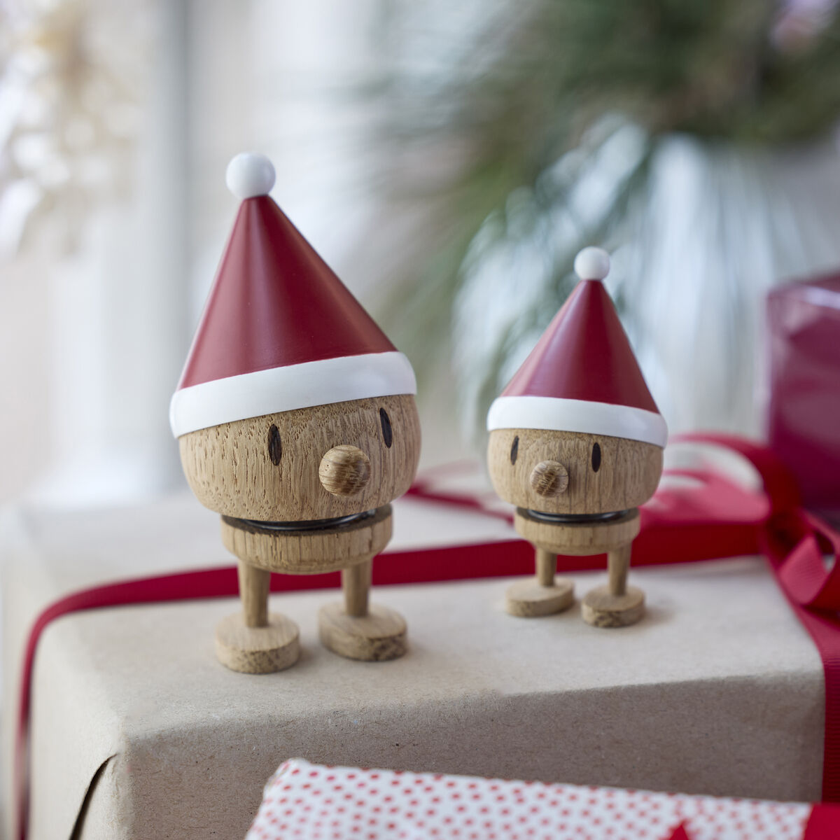 Zwei Hoptimist Elf Bumble Raw oak Holzfiguren mit roten Weihnachtsmützen stehen auf weihnachtlich verpackten Geschenken mit roten Schleifen. Im Hintergrund ist unscharfe Dekoration und eine Pflanze zu erkennen. Die Szene wirkt festlich und gemütlich.