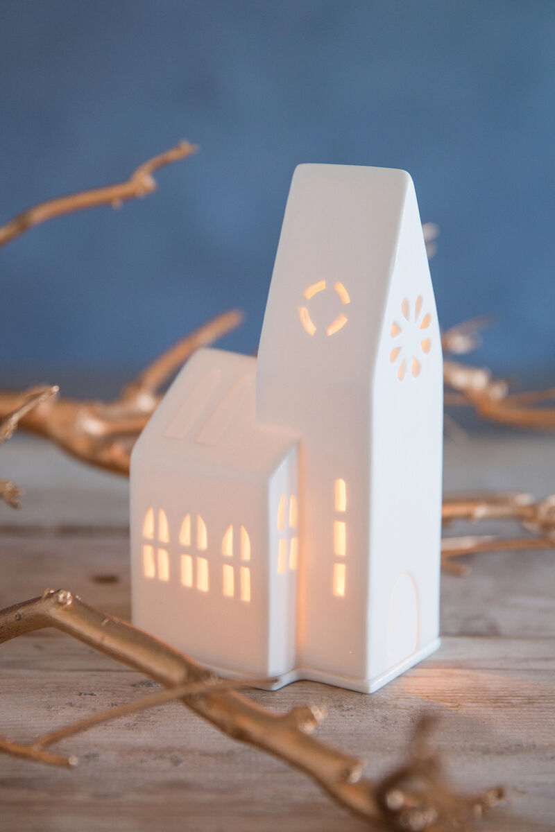 Keramik-Lichthaus in Form einer kleinen Kirche mit leuchtenden Fenstern und einer stilisierten Glocke, umgeben von goldenen Ästen auf einem Holztisch.