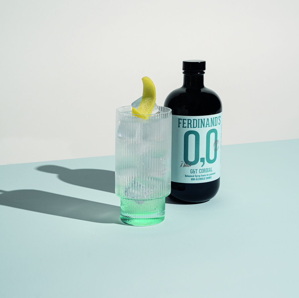 Ferdinand's 0,0 G&T Cordial-Flasche neben einem Glas mit transparentem Getränk und Zitronengarnitur auf einer türkisen Oberfläche.