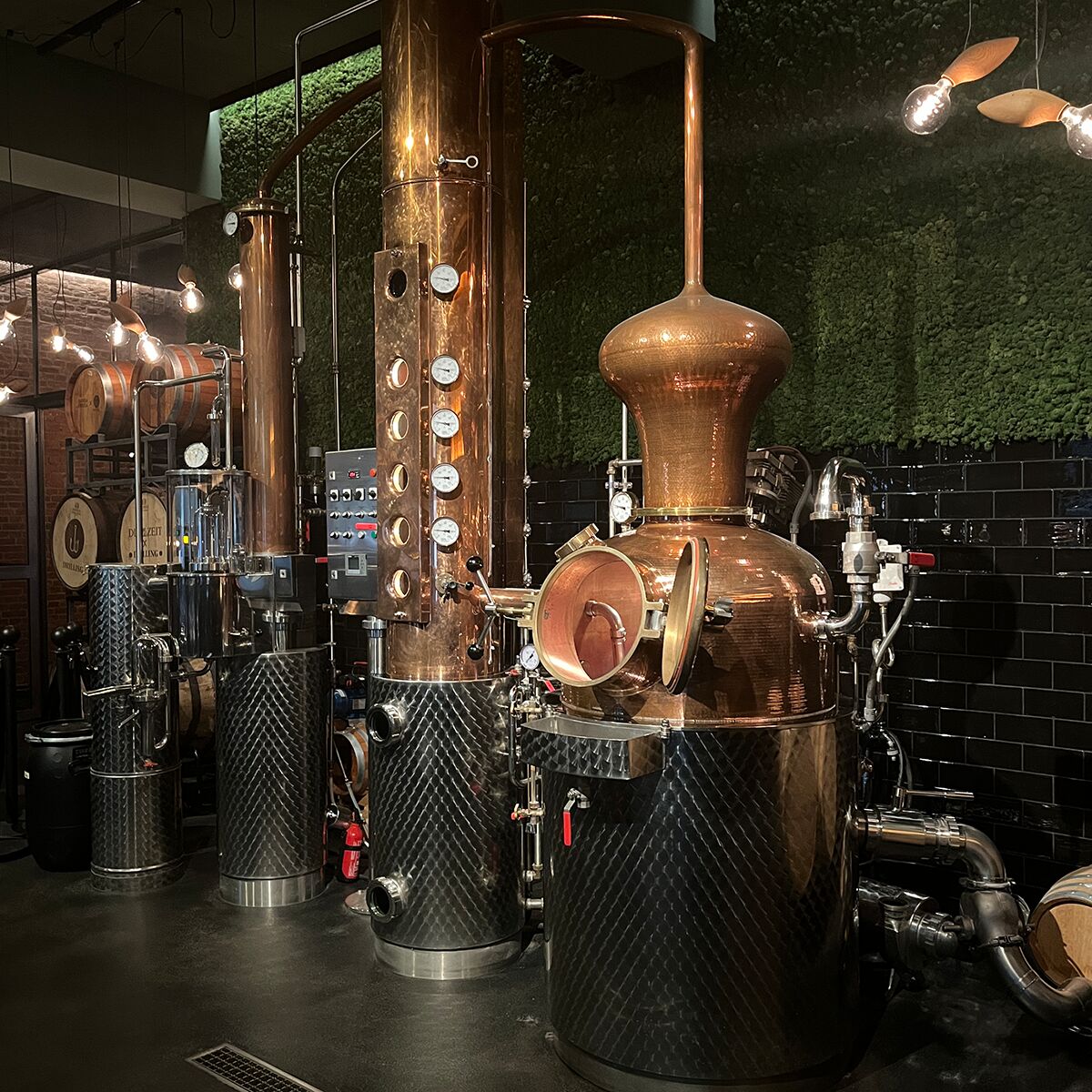 Eine moderne Destillerie mit glänzenden kupfernen Brennblasen und subtilen, schwarzen und grünen Wandakzenten. Die Anlage des Single Malt Whiskey »Drilling« der ZEIT-Sonderedition steht im Vordergrund, umgeben von handwerklichen Details und hochwertigen Materialien. Die Szene vermittelt einen Eindruck von Qualität, Tradition und exklusivem Genuss.