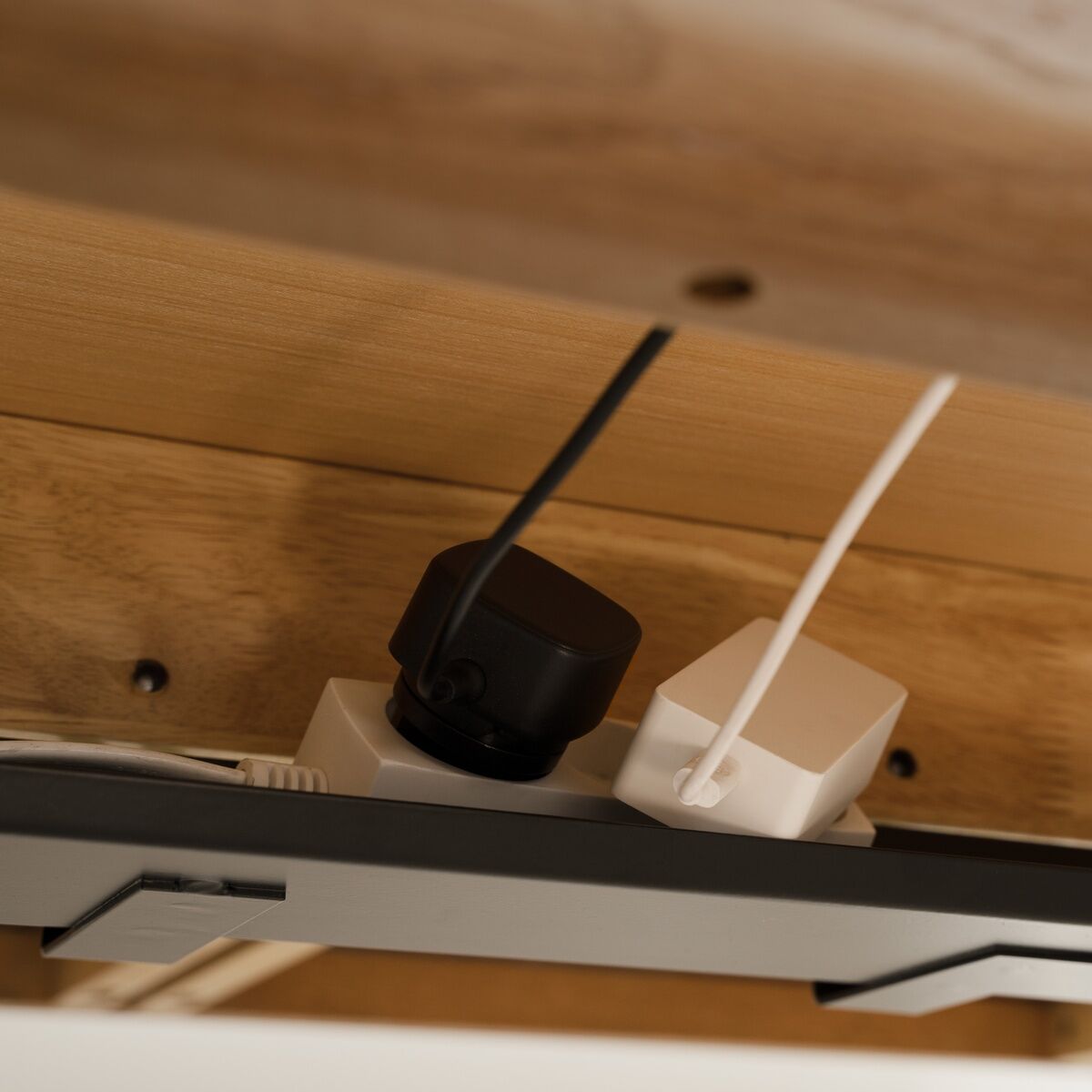 Detailansicht unter einer Schreibtischplatte aus Holz. Eine Steckdosenleiste mit weißen und schwarzen Steckern ist ordentlich an der Unterseite des Schreibtischs »Heart'n'Soul« der Marke Umage montiert, Kabel verlaufen geordnet entlang der Führung.