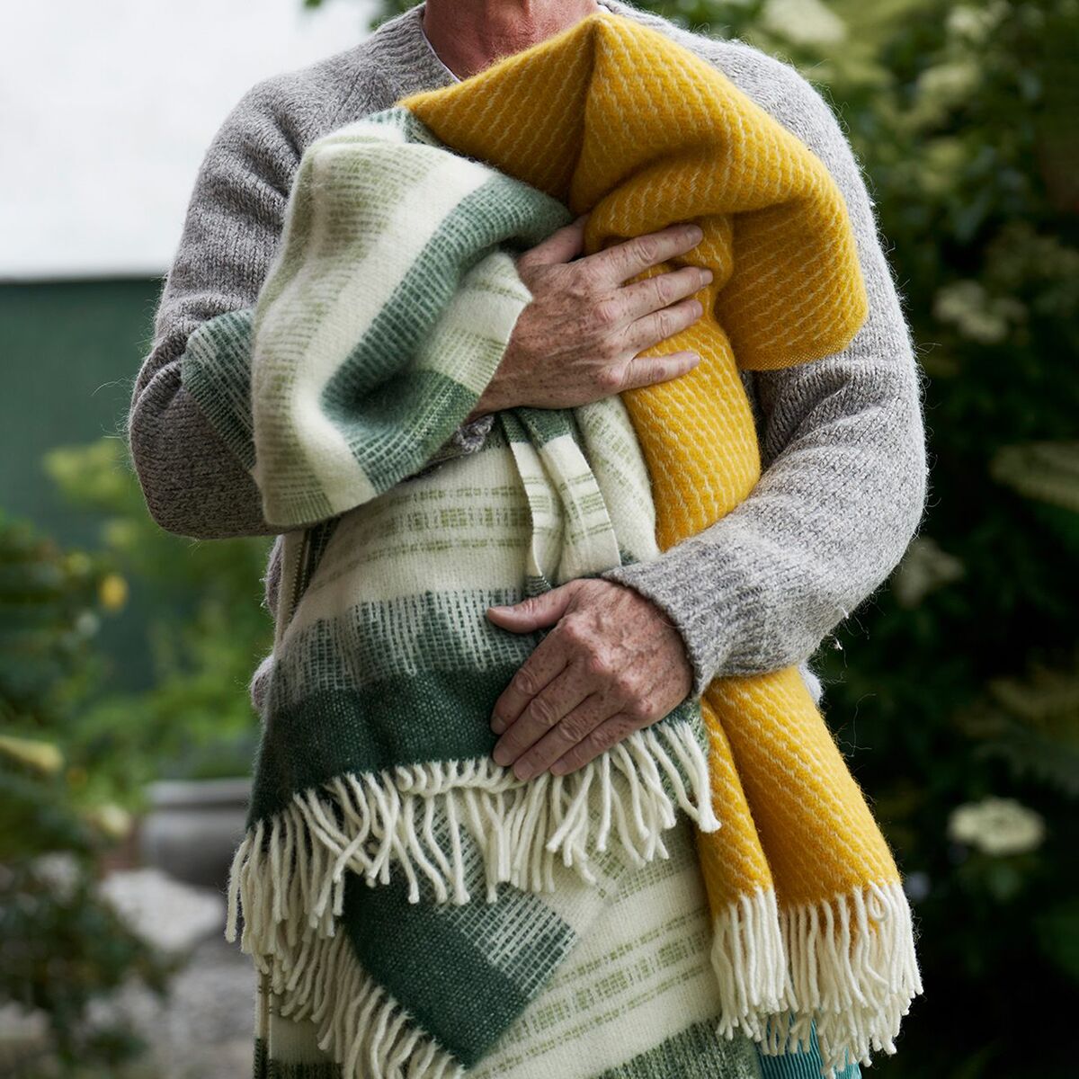 Person hält zwei weiche, gemusterte Decken in Gelb, Grün, Weiß und Creme, zusammengeschlungen vor einem Gartenhintergrund. Die Szene vermittelt Gemütlichkeit und Wärme.