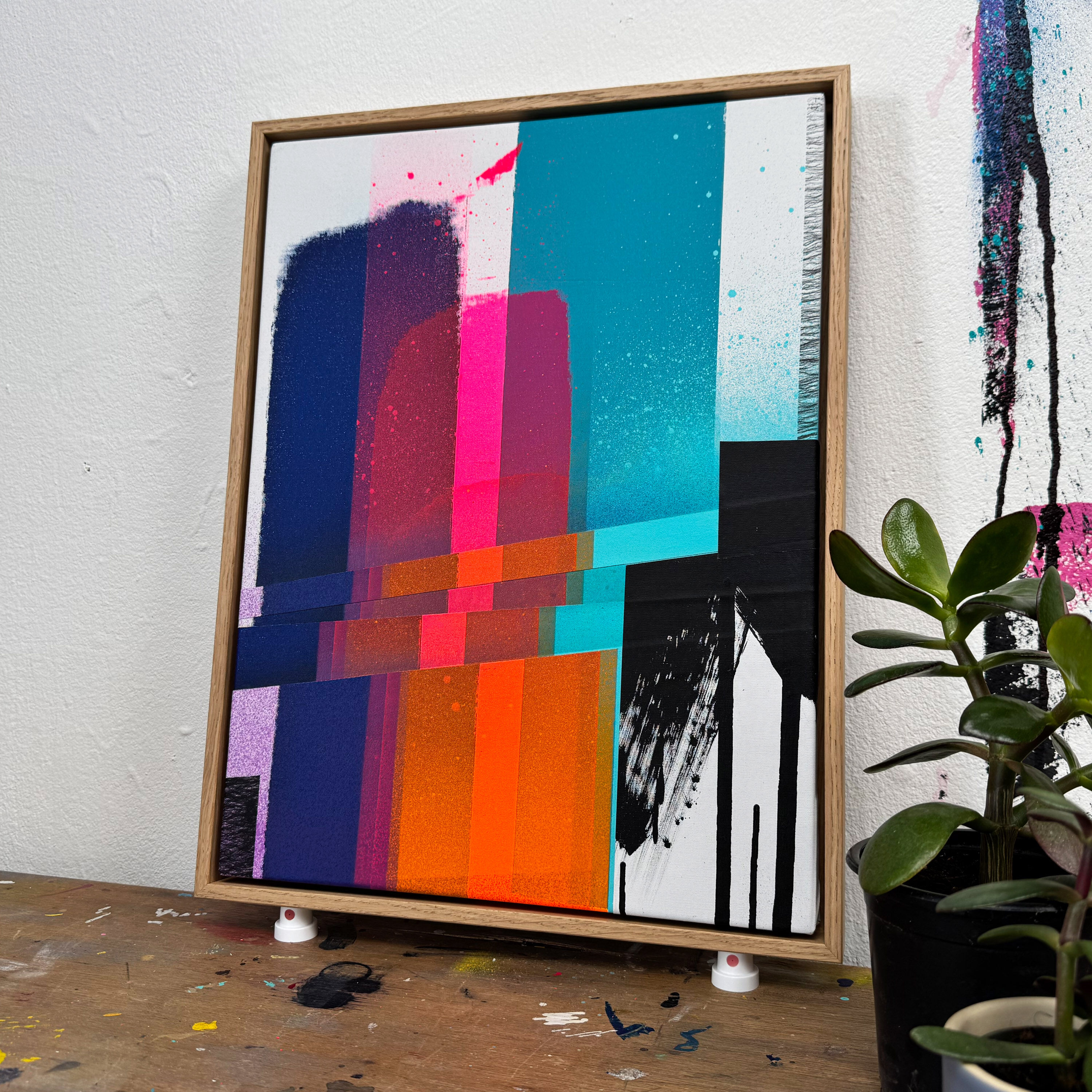 Farbenfrohes, abstraktes Siebdruck-Unikat mit dynamischen geometrischen Formen in Pink, Blau, Orange, Türkis, Schwarz und Weiß, eingerahmt und auf einem Holztisch stehend, neben einer Zimmerpflanze, vor einer weißen Wand.