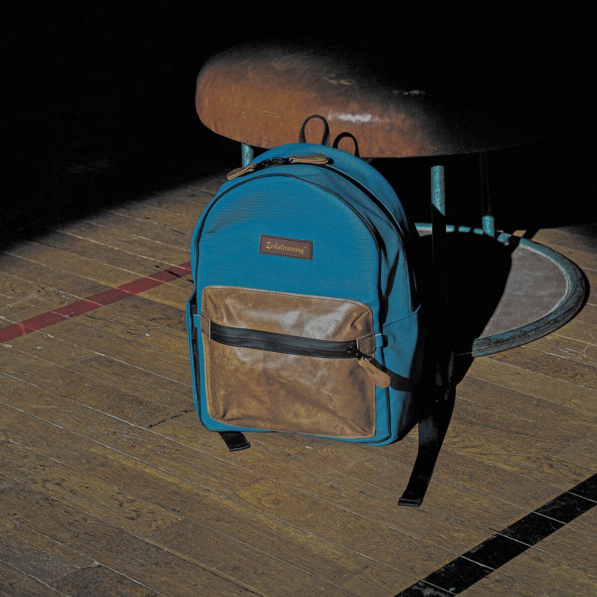 Ein türkisfarbener Rucksack mit braunen Lederapplikationen steht unter einem Lichtstrahl auf einem Holzboden. Im Hintergrund ist teilweise eine Gymnastikbank zu sehen, die im Schatten liegt.