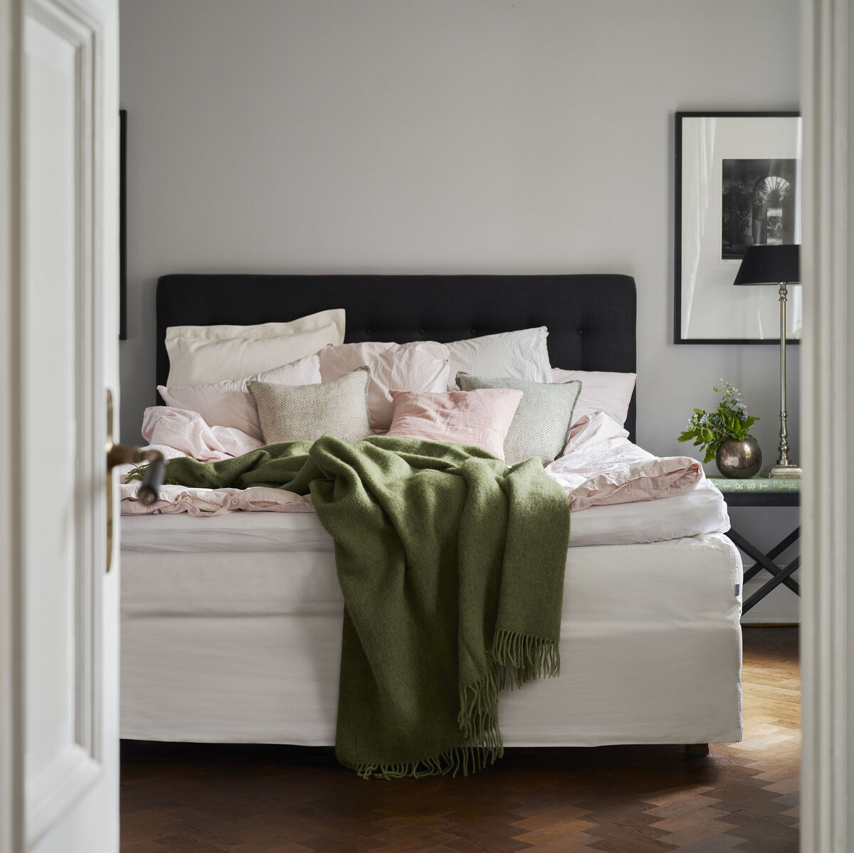 Schlafzimmeransicht mit Türrahmen, weißen Wänden und einem gemütlichen Bett. Über dem Bett hängt mit grüner Decke Gotland und mehreren Kissen. Holzfußboden sowie Kunstwerke und eine Pflanze im Hintergrund ergänzen die Inneneinrichtung.