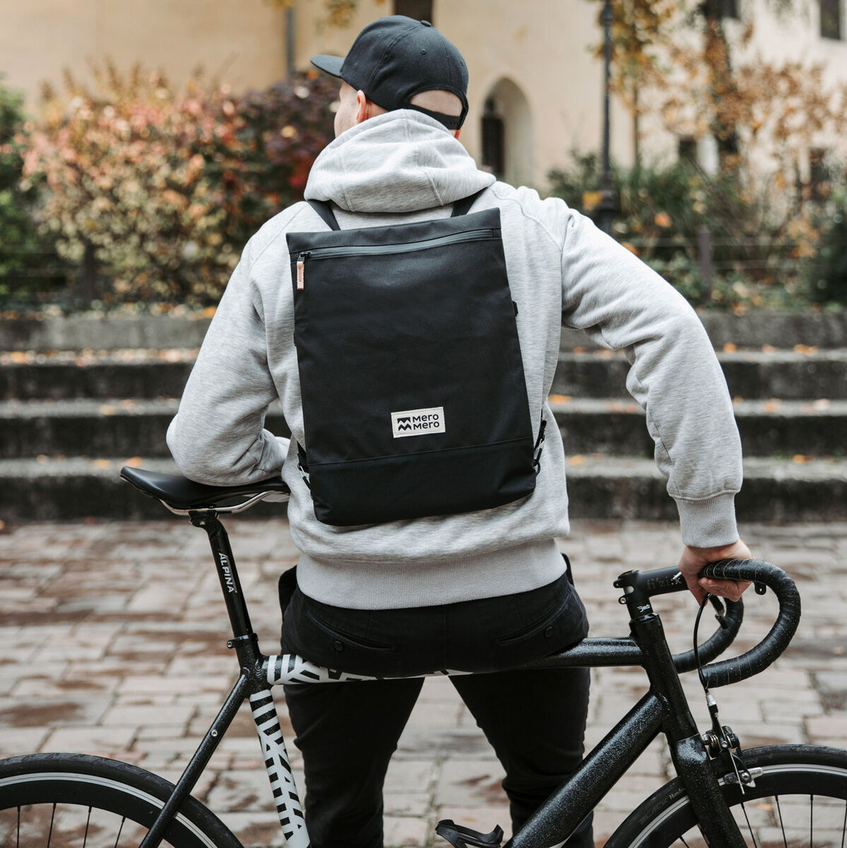 Eine Person mit einer grauen Kapuzenjacke und einem schwarzen Pouch »Meije«-Rucksack auf dem Rücken schiebt ein Fahrrad. Sie steht in einer herbstlichen, städtischen Umgebung mit Bäumen und einem Gebäude im Hintergrund.