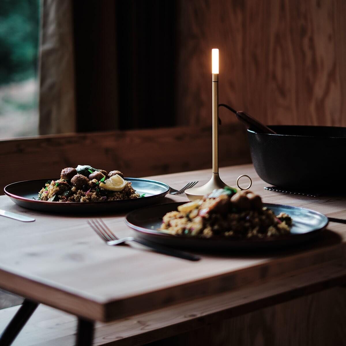 Ein stilvoll gedeckter Holztisch mit zwei Tellern, gefüllt mit Essen. Auf dem Tisch steht eine schlanke, moderne, goldene Wick Tischlampe, die ein sanftes, warmes Licht abgibt. Im Hintergrund befindet sich eine große, schwarze Pfanne.