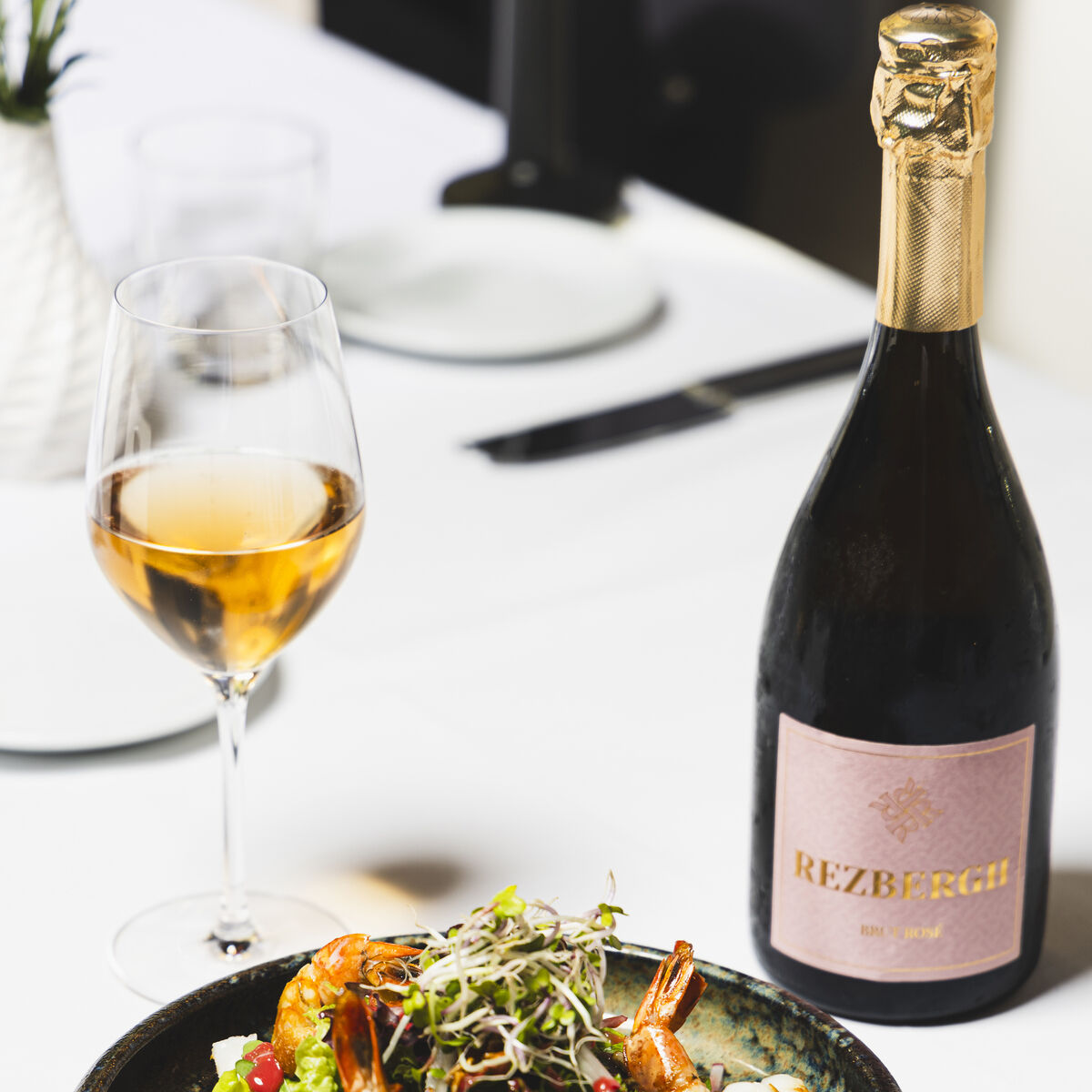 Eine Flasche Crémant de Luxembourg Brut Blanc des Blancs Réserve steht auf einem weiß gedeckten Tisch neben einem gefüllten Weinglas und einer eleganten Schale mit Salat und Garnelen. Das Arrangement vermittelt einen hochwertigen und festlichen Eindruck, geeignet für besondere Anlässe oder Feiern.