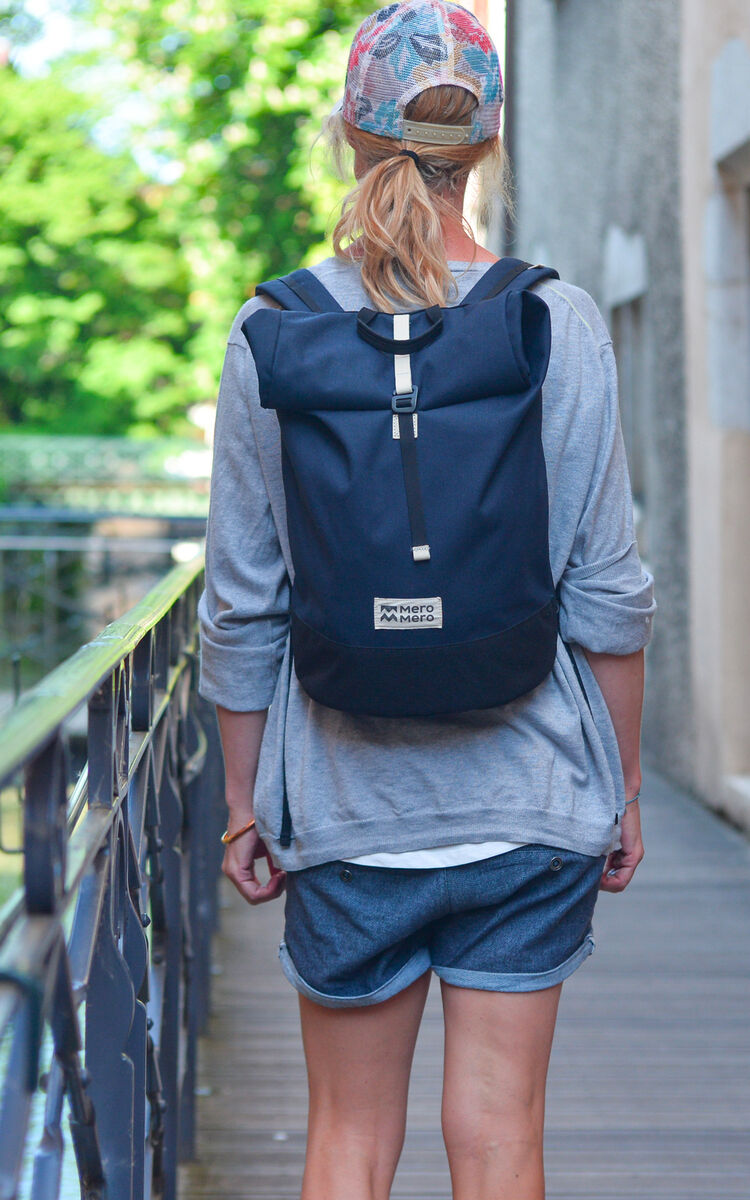 Person trägt blaue Fahrradtasche »Annecy« auf dem Rücken. Der Hintergrund zeigt eine städtische Umgebung mit Bäumen und Gebäuden.