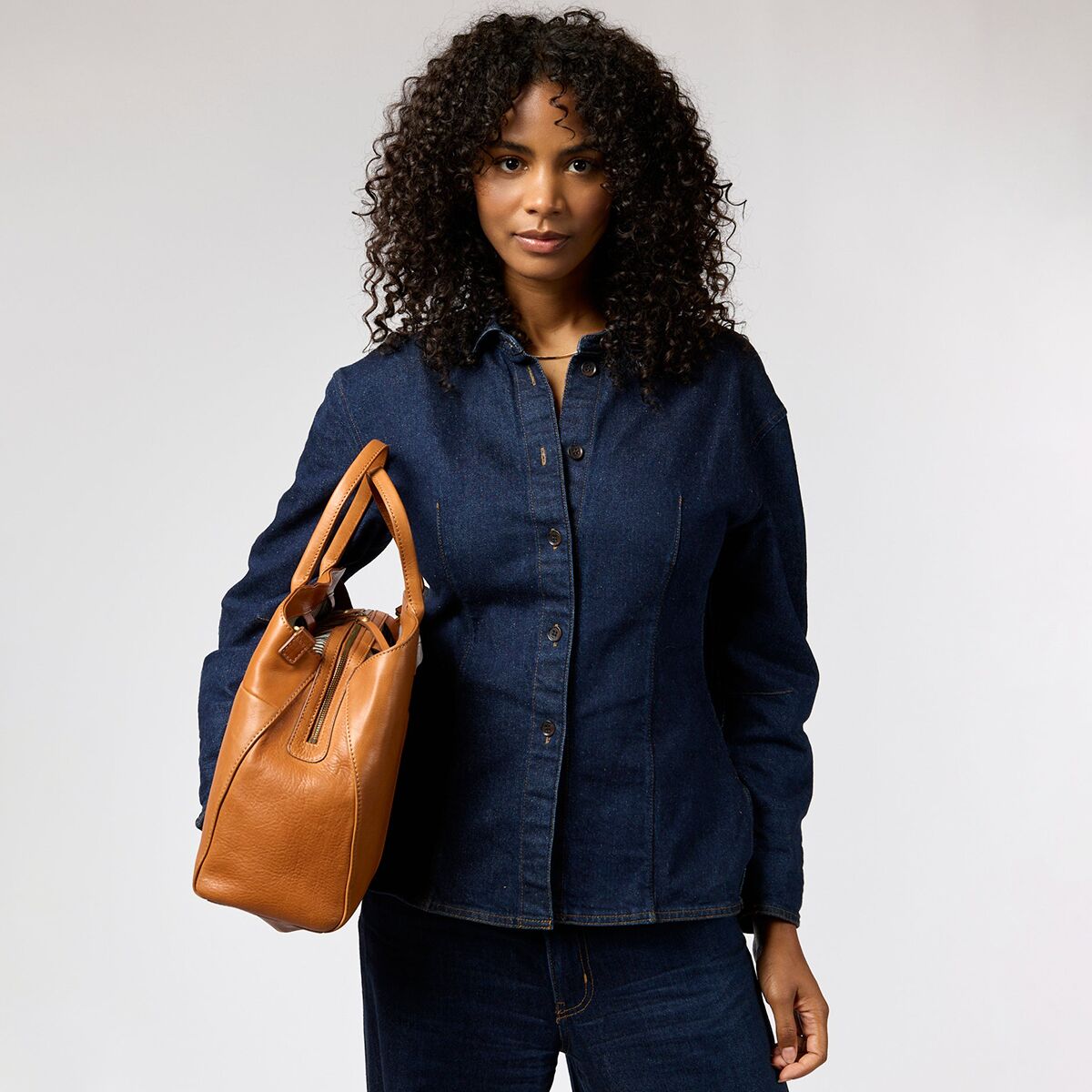 Eine Frau trägt eine mittelgroße, braune Schultertasche aus Narbenleder der Marke O My Bag Modell Rita am rechten Arm. Sie trägt eine dunkelblaue Jeansjacke und dazu passende Jeans. Die Tasche hat goldene Details und eine strukturierte Oberfläche. Der Hintergrund ist neutral weiß.