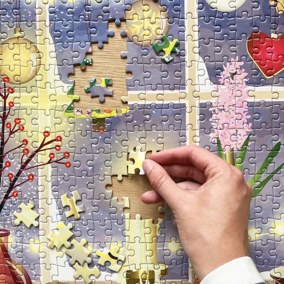 Nahaufnahme eines sich in Fertigstellung befindenden Puzzles mit 500 Teilen von VISSEVASSE. Das Motiv zeigt ein festlich dekoriertes Fenster mit Kerze, Weihnachtsbaum, Kugeln, Herzanhänger sowie Blumen. Eine Hand setzt gerade ein Puzzleteil ein.