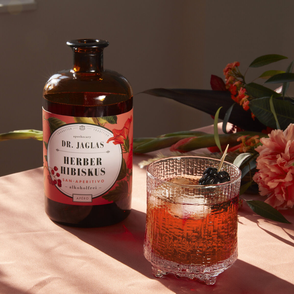 Eine Flasche 'Herber Hibiskus' alkoholfreier Aperitif von Dr. Jaglas steht auf einem Tisch neben einem Kristallglas mit dem Getränk, dekoriert mit Eiswürfeln und Beeren.
