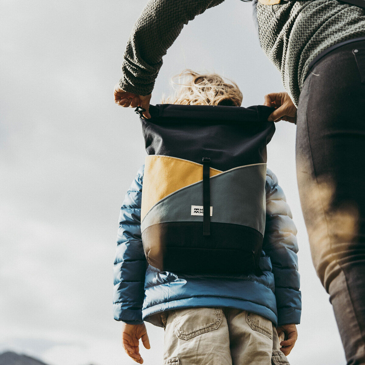 Person hält einen Rucksack »Popoyo« im Freien. Der Rucksack ist mehrfarbig, hauptsächlich in Schwarz, Grau, Gelb, Blau und etwas Braun. Der Hintergrund zeigt eine natürliche Landschaft mit Wolken und Himmel.