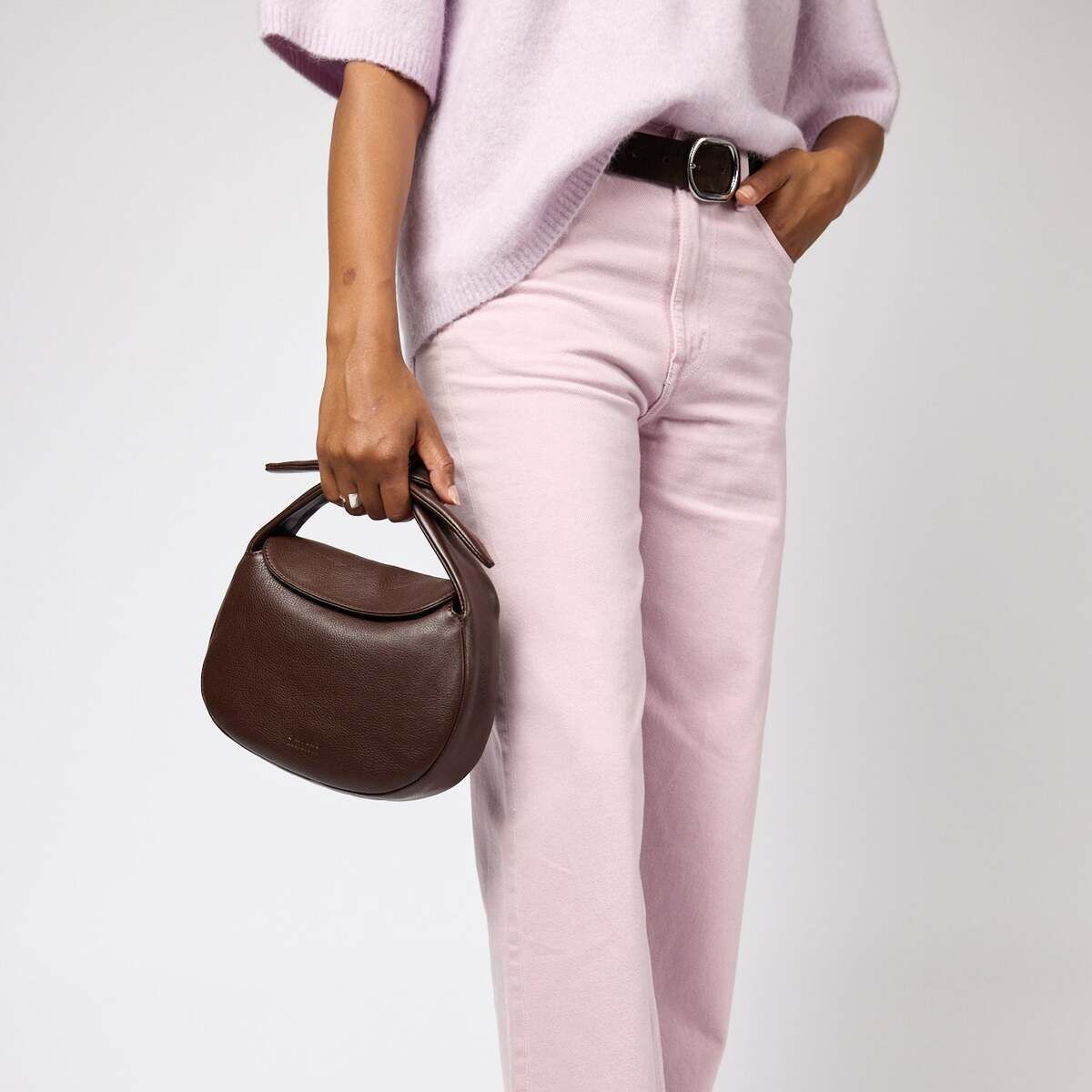 Frau hält eine dunkelbraune Handtasche »Coco Croissant« in der linken Hand, trägt hellrosa Jeans, braune spitze Schuhe und ein fliederfarbenes Oberteil vor klarem Hintergrund.
