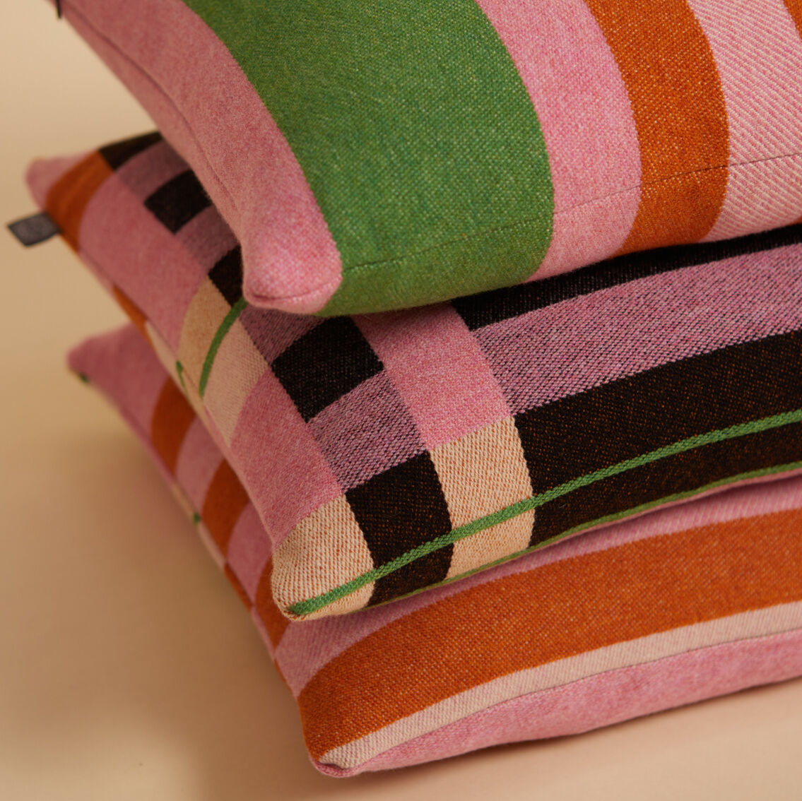 Ein Stapel von drei dekorativen Kissen mit einem geometrischen Streifenmuster in Rosa, Grün, Orange, Schwarz und Weiß. Das Design der Kissen erinnert an den Bauhaus-Stil.