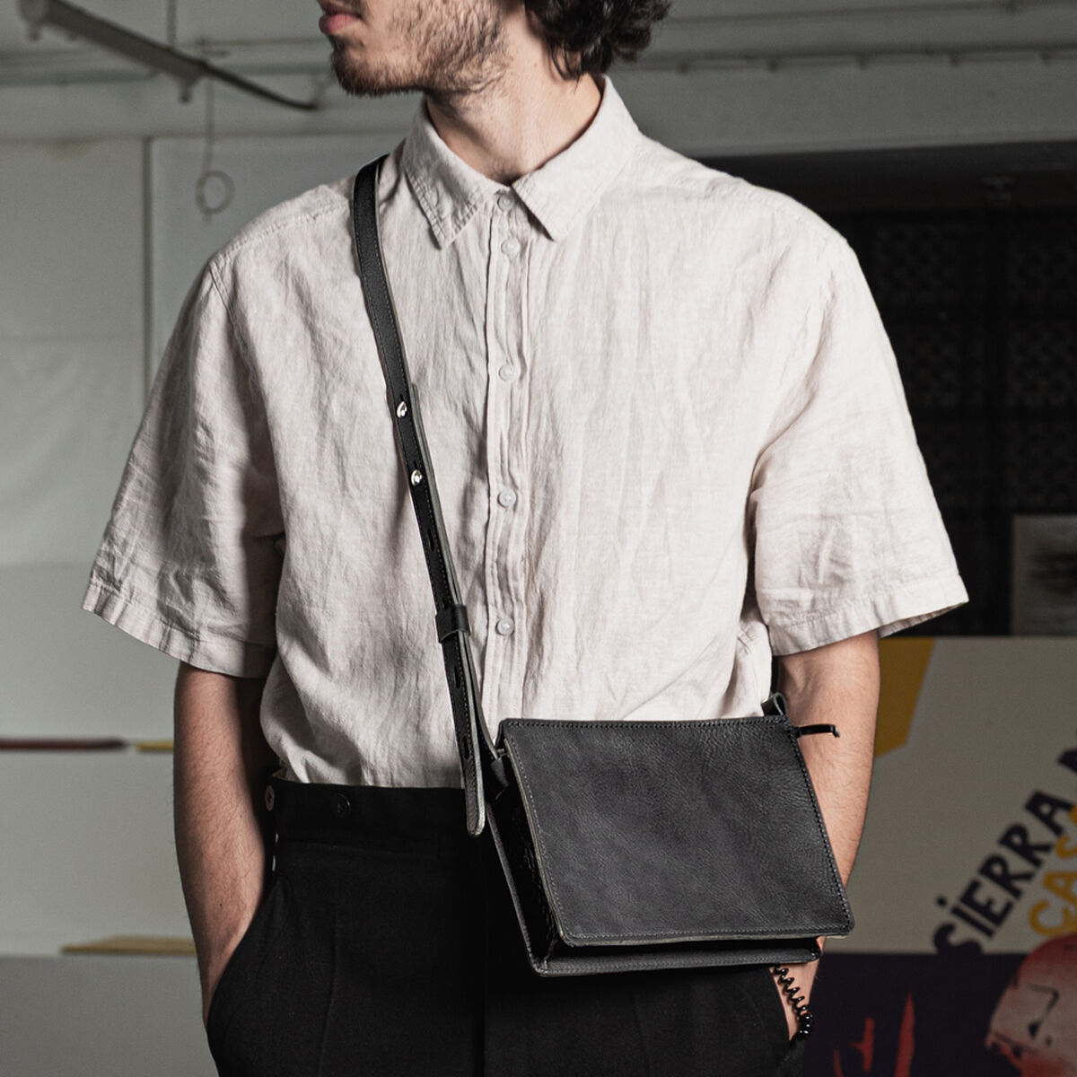 Mann trägt eine schwarze Kernleder-Umhängetasche »Sas« über der Schulter. Er trägt ein helles, kurzärmeliges Hemd und dunkle Hose. Die hochwertige Ledertasche ist minimalistisch gestaltet und eignet sich für den modernen urbanen Alltag.