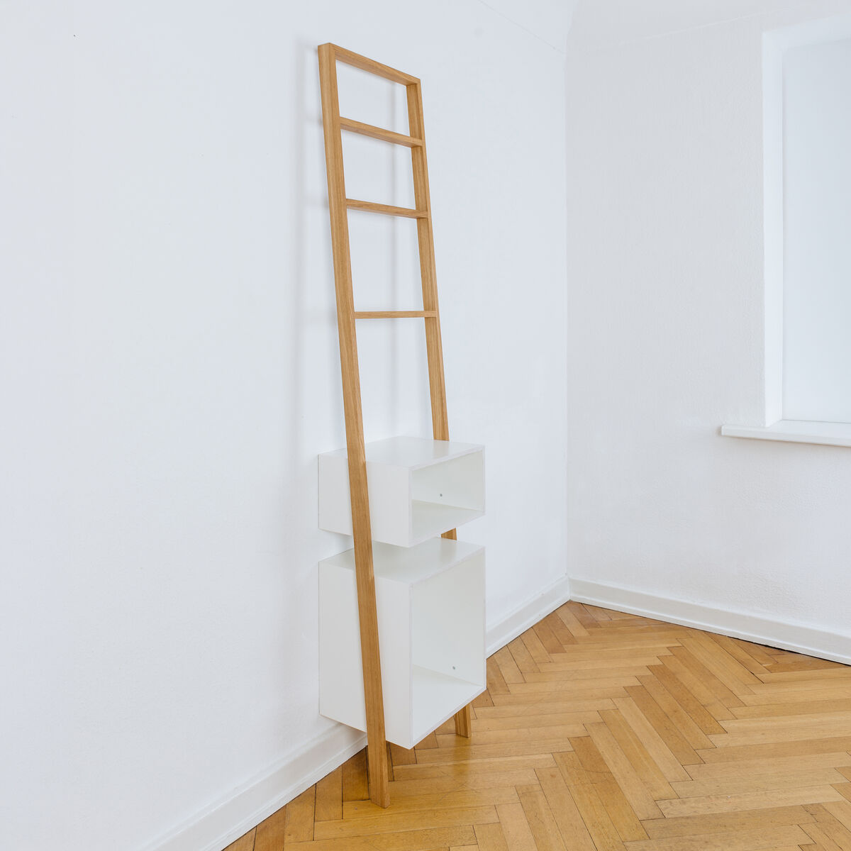 Modernes Leiterregal aus geölter Eiche mit zwei weißen Regalkästen steht an einer weißen Wand auf hellem Fischgrätparkett. Das Möbelstück überzeugt durch schlichtes, minimalistisches Design und eignet sich zur Aufbewahrung von Büchern oder Deko in kleinen Räumen.