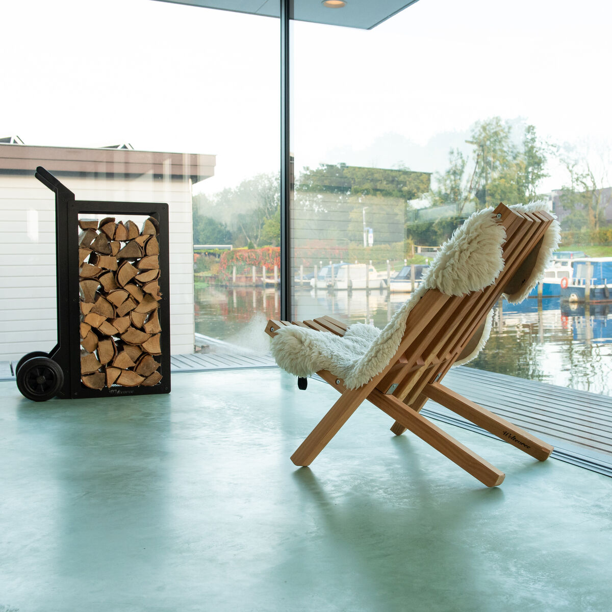 Ein stilvoller Holzstuhl mit Schafsfellauflage steht in einem modernen, lichtdurchfluteten Raum mit Blick auf einen ruhigen See. Daneben ein Holzstapel auf einem Rollwagen. Perfektes Ambiente für Entspannung.
