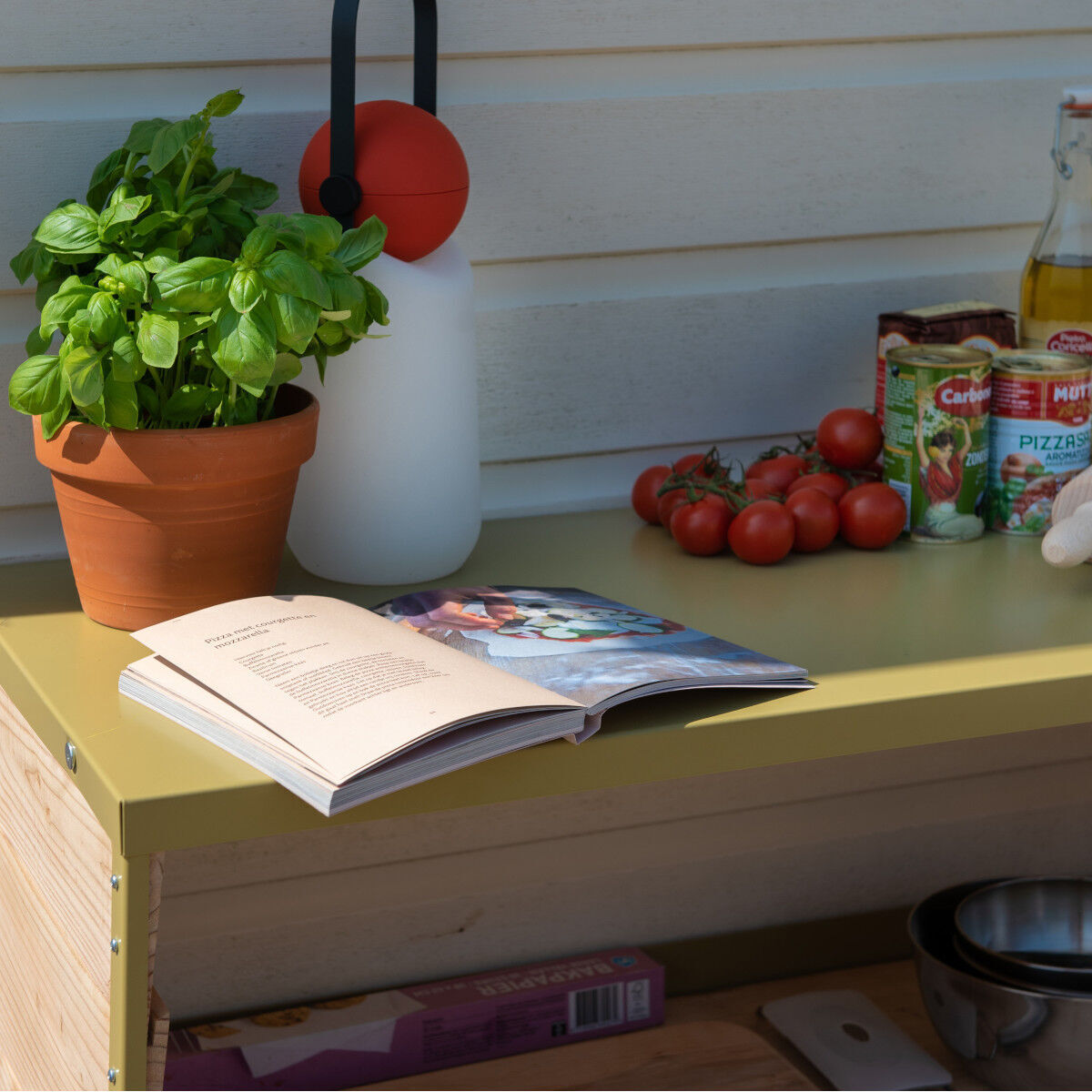 Eine weiße Guidelight Lampe mit einem roten Griff steht neben einem Topf Basilikum, einer Flasche Öl, einer Pizza-Tomatendose und frischen Tomaten auf einem grünen Tisch. Ein offenes Kochbuch liegt ebenfalls auf dem Tisch.