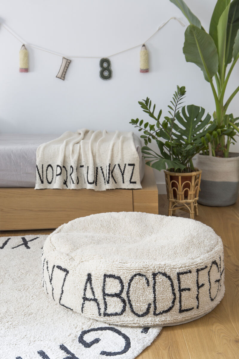 Ein rundes Sitzkissen »Pouf ABC« aus weißem Stoff mit schwarzen Buchstaben des Alphabets darauf. Es befindet sich in einem modern eingerichteten Zimmer mit einer Pflanze im Hintergrund und einem Teppich, der ebenfalls mit Buchstaben bedruckt ist.