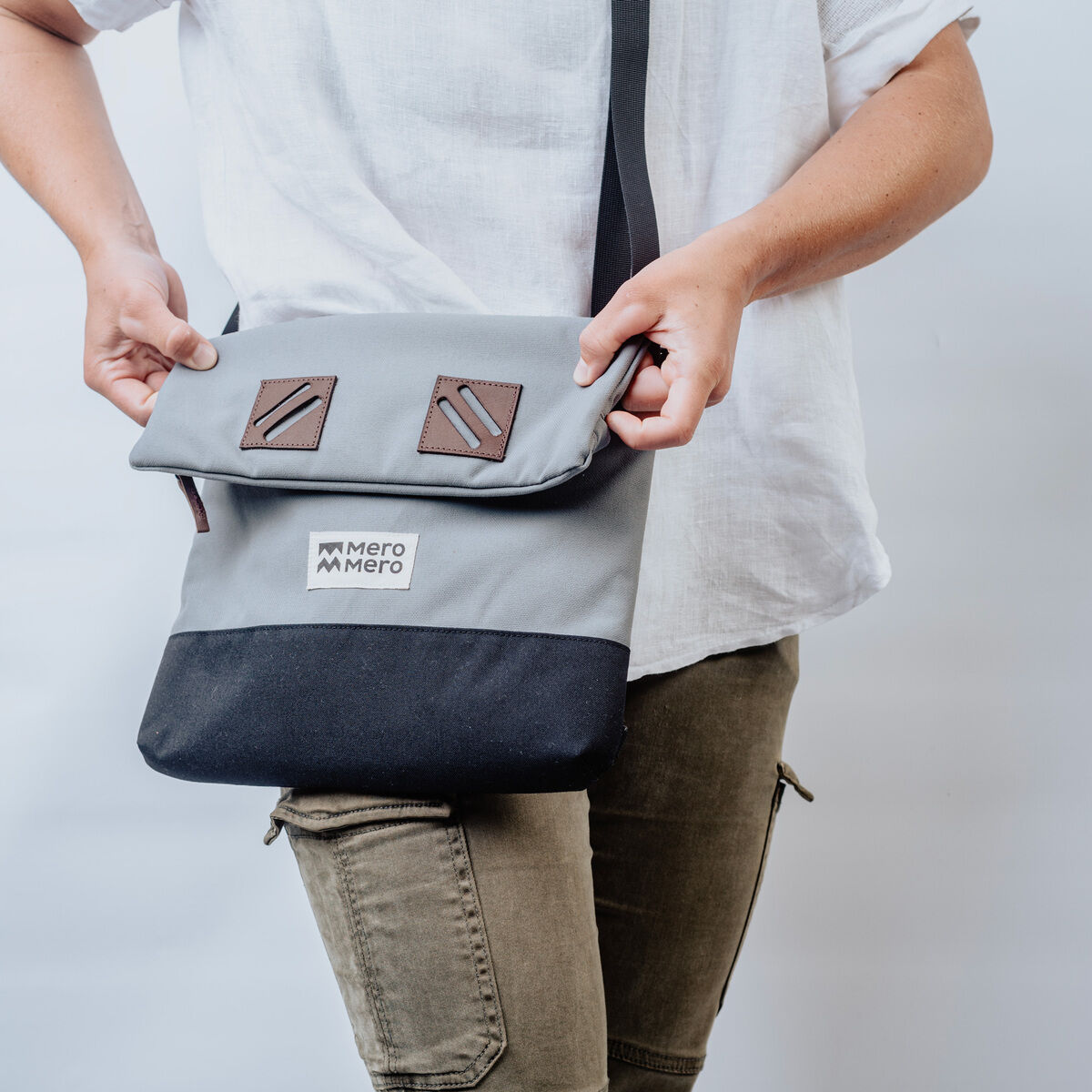 Person trägt eine graue und schwarze Tasche »Meije« von mero mero über der Schulter. Die Tasche ist schlicht und minimalistisch gestaltet mit zwei braunen Zierdetails und einem weißen Logo auf der Vorderseite.