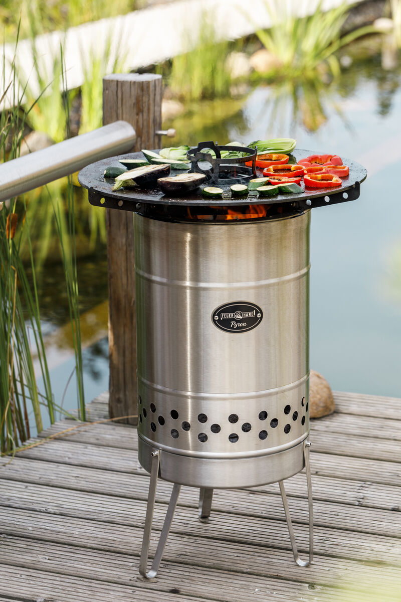 Eine Feuertonne aus Edelstahl der Marke »Pyron« steht auf einer Holzterrasse. Auf dem Grillrost oberhalb der Tonne befinden sich Scheiben von Zucchini und Paprika. Im Hintergrund ist ein Teich mit Schilf und Gras zu sehen, der eine entspannte Outdoor-Atmosphäre vermittelt.
